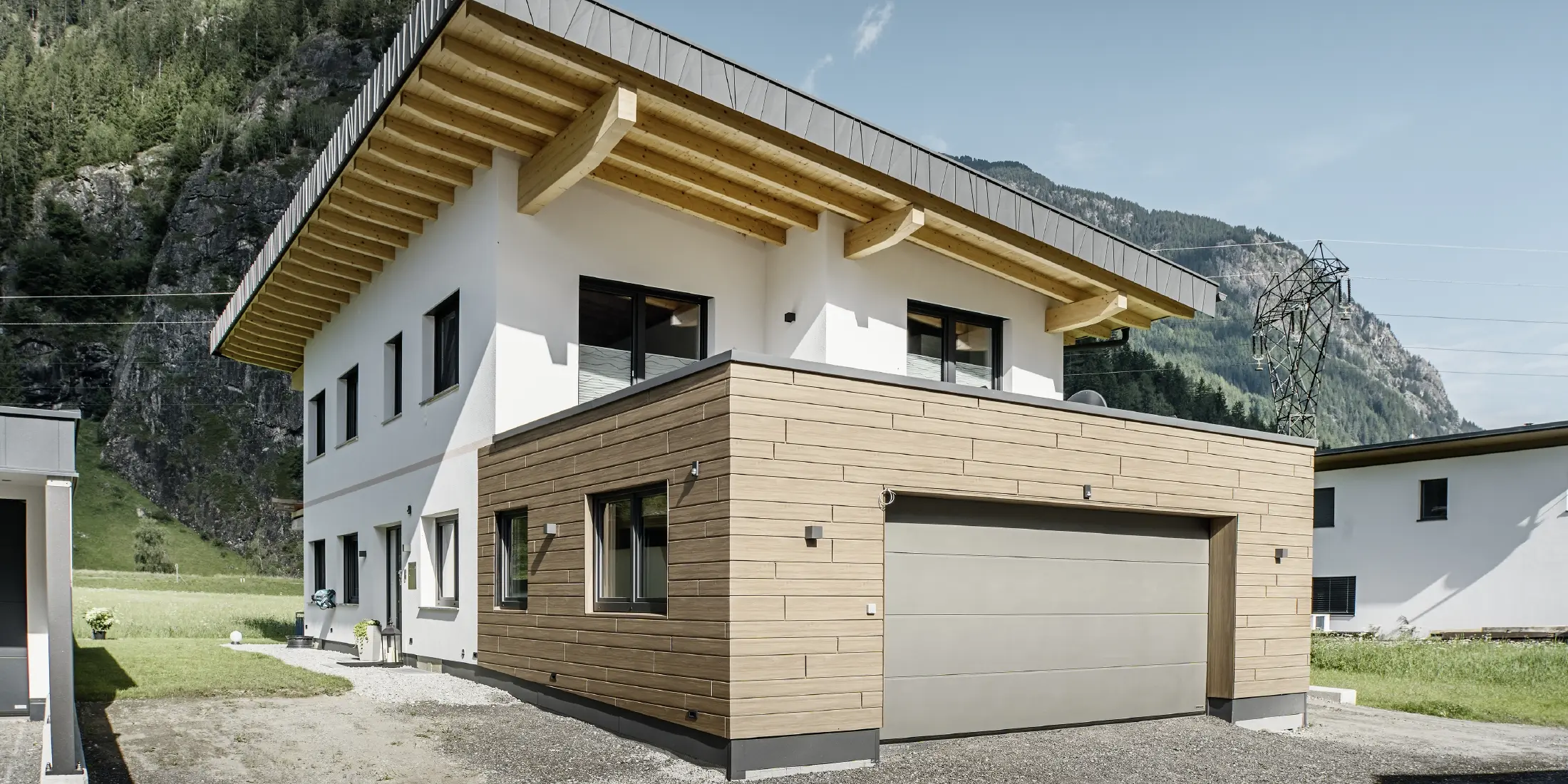 Moderne eengezinswoning met gevelrenovatie: vernieuwde buitengevel met horizontaal geplaatste gevelbekleding in houtlook, gecombineerd met wit gepleisterde vlakken, geïntegreerde garagebekleding en eigentijdse architectuur in een alpiene omgeving.