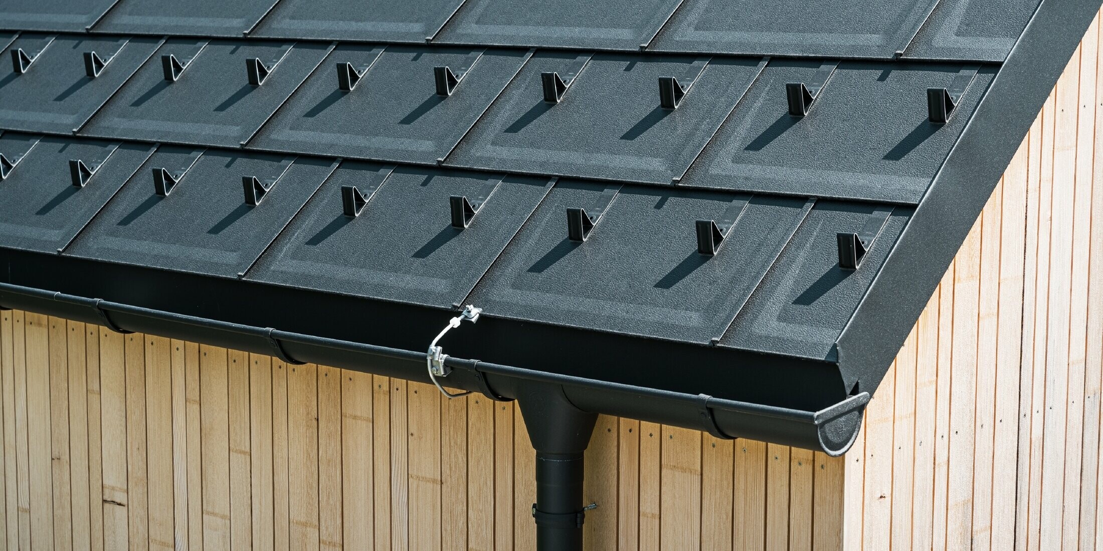 Detailafbeelding van de PREFA dakplaat R.16 met sneeuwstop, dakgoot en afvoerbuis in zwart P.10 incl. bliksembeveiliging en een verticale houten gevel