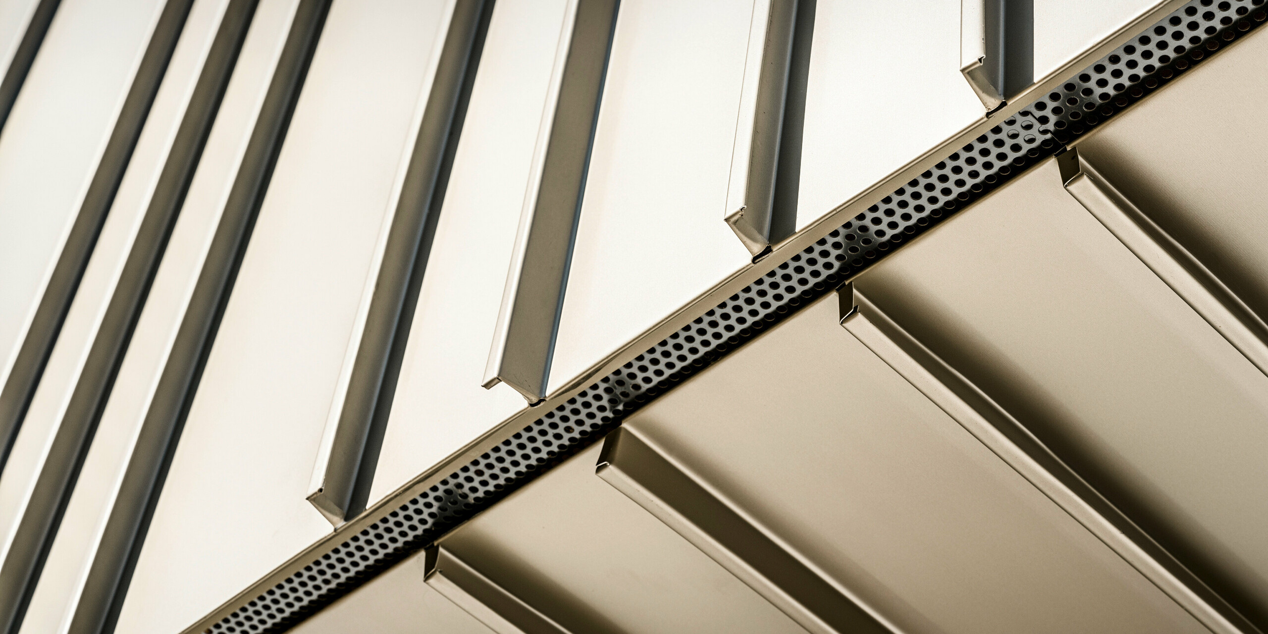 Detailaufnahme einer innovativen PREFA Aluminiumfassade in der Farbe Lichtbronze. Die vertikale Winkelstehfalzdeckung und die präzise Kantenführung unterstreichen die hervorragende Verarbeitung. Die sichtbaren Perforationen sorgen für eine moderne Optik und bieten zudem auch funktionale Vorteile, wie verbesserte Belüftung. Diese Komponenten spiegeln die technische Raffinesse und das ökologische Bewusstsein von Aluminium als Baumaterial wider.