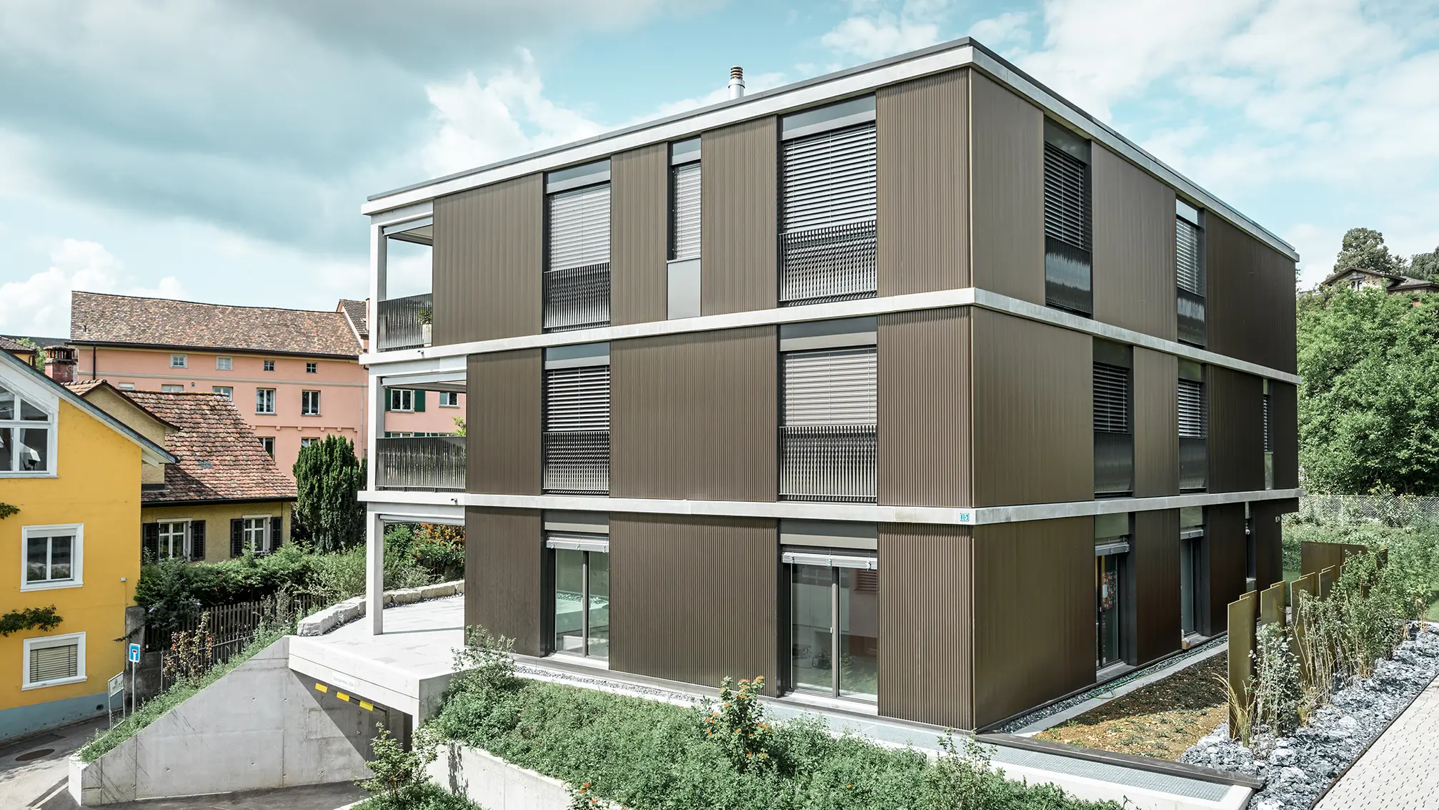 De opvallende aluminium gevel van dit appartementencomplex in Schaffhausen is voorzien van het elegante PREFA-kartelprofiel in de kleur Colinal. Door de verticale plaatsing van de geëxtrudeerde aluminium profielen oogt het gebouw optisch compacter en krijgt de gevelstructuur een harmonieuze balans. Het elegante ontwerp past smaakvol in de omgeving en geeft het gebouw een moderne elegantie die zowel visueel aantrekkelijk als functioneel is. Het perspectief toont het appartementencomplex vanaf de voorzijde met de garage-ingang. Op de achtergrond zijn bomen, andere gebouwen en een kerktoren te zien.