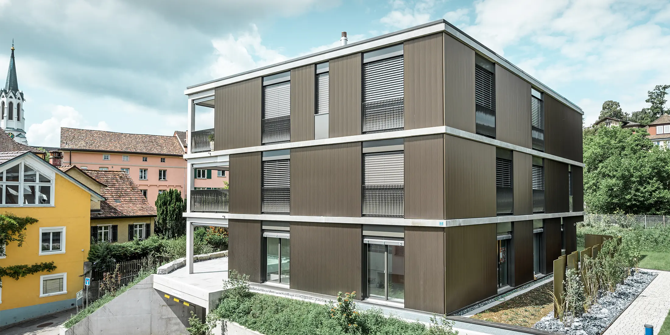 De opvallende aluminium gevel van dit appartementencomplex in Schaffhausen is voorzien van het elegante PREFA-kartelprofiel in de kleur Colinal. Door de verticale plaatsing van de geëxtrudeerde aluminium profielen oogt het gebouw optisch compacter en krijgt de gevelstructuur een harmonieuze balans. Het elegante ontwerp past smaakvol in de omgeving en geeft het gebouw een moderne elegantie die zowel visueel aantrekkelijk als functioneel is. Het perspectief toont het appartementencomplex vanaf de voorzijde met de garage-ingang. Op de achtergrond zijn bomen, andere gebouwen en een kerktoren te zien.