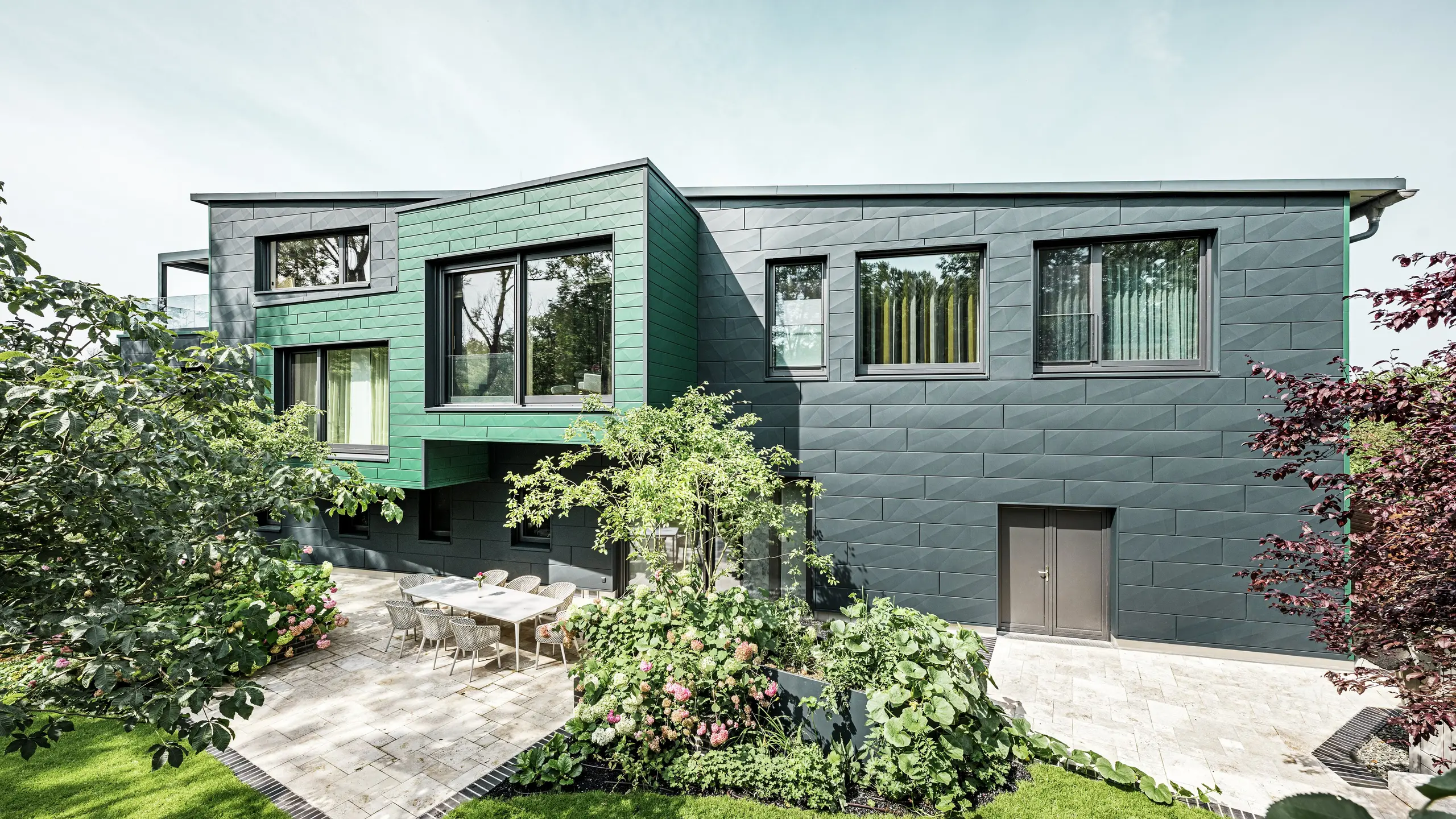 De eengezinswoning in Türkheim maakt indruk met zijn bijzondere gevel, die is bekleed met PREFA Siding X-panelen in de kleuren antraciet en mosgroen. De strakke lijnen en de opvallende kleurkeuze van de aluminium panelen geven het gebouw een moderne en elegante uitstraling. Op de voorgrond is de royale tuin te zien met een stijlvol terras en een eettafel met moderne stoelen. Kleurrijke bloembedden en gezonde planten zorgen voor een levendig, groen contrast met de robuuste gevel. De duurzame en onderhoudsvriendelijke aluminium gevel beschermt het gebouw en zorgt voor tijdloze schoonheid.
