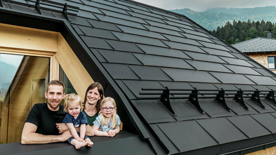De twee opdrachtgevers genieten samen met hun kinderen van het uitzicht vanaf het inpandige balkon van hun nieuwbouwwoning, die is voorzien van stijlvolle PREFA-solar-dakpannen in P.10 zwart.