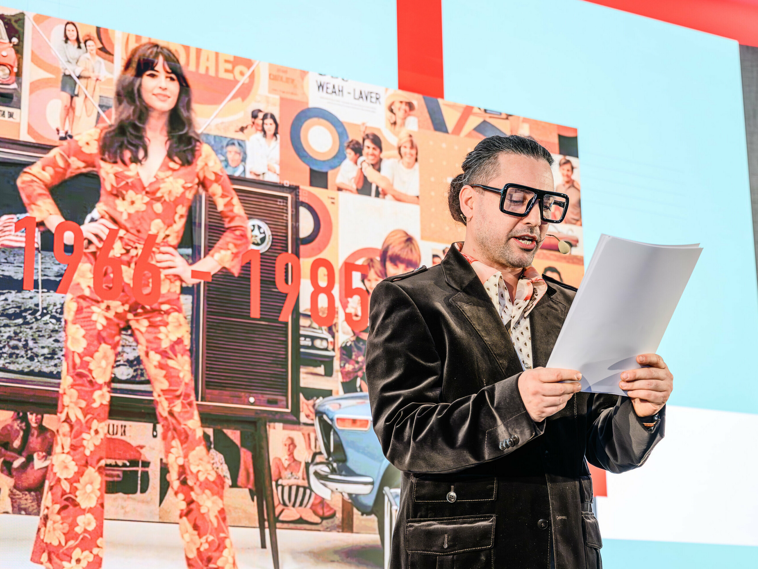 Zeitreise auf der Bühne der PREFARENZEN-Präsentation 2026: Der charismatische Serkan Temel im Retro-Look führt das Publikum durch die Jahrzehnte. Im Fokus stehen die Jahre 1966 bis 1985, die auf einer Collage im Hintergrund mit Design-Ikonen und gesellschaftlichen Trends dieser Ära dargestellt sind. Genau wie PREFA-Produkte überdauern Trends mit Stil und Substanz. Die Präsentation zeigt, wie Architektur von gestern Inspiration für die Lösungen von morgen wird – mit Aluminium neu interpretiert.