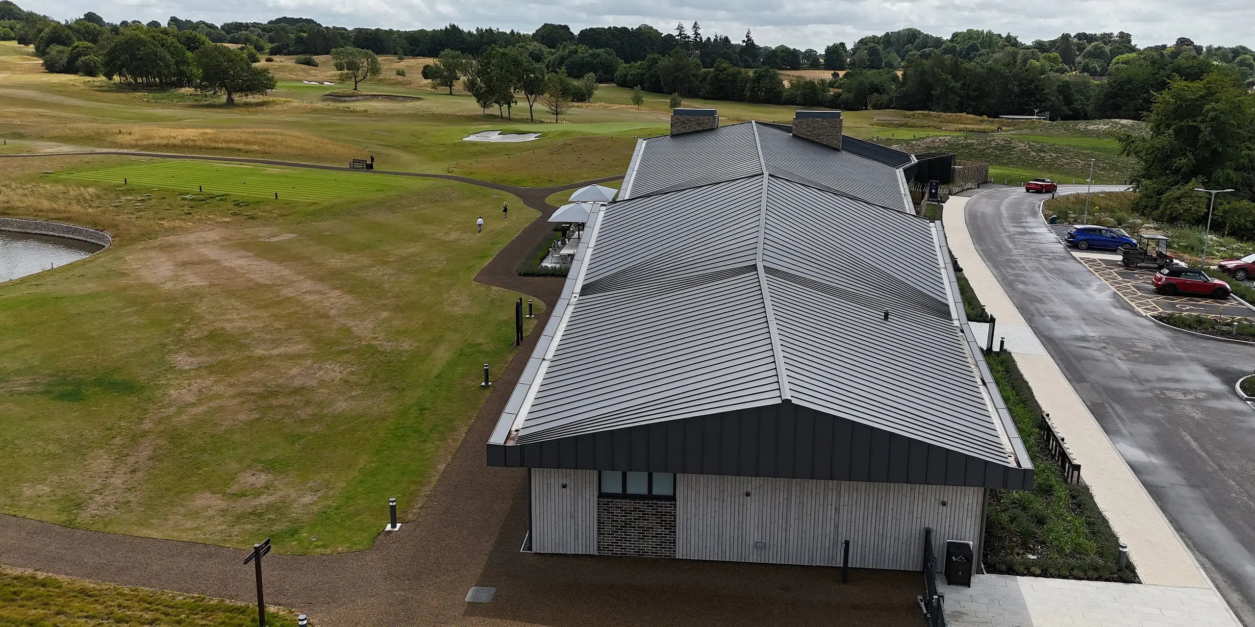 Das elegante PREFALZ Stehfalz-Dach des Basingstoke Golf Clubs in Hampshire zieht mit seiner geschwungenen Form alle Blicke auf sich. Das Dach in P.10 Dunkelgrau setzt markante Akzente, die sich harmonisch in die weite Golfplatzlandschaft einfügen. Im Vordergrund ist die Giebelseite des Gebäudes sowie das gepflegte Gelände des Golfplatzes zu sehen, das sich ideal mit der modernen Architektur des Clubhauses vereint.