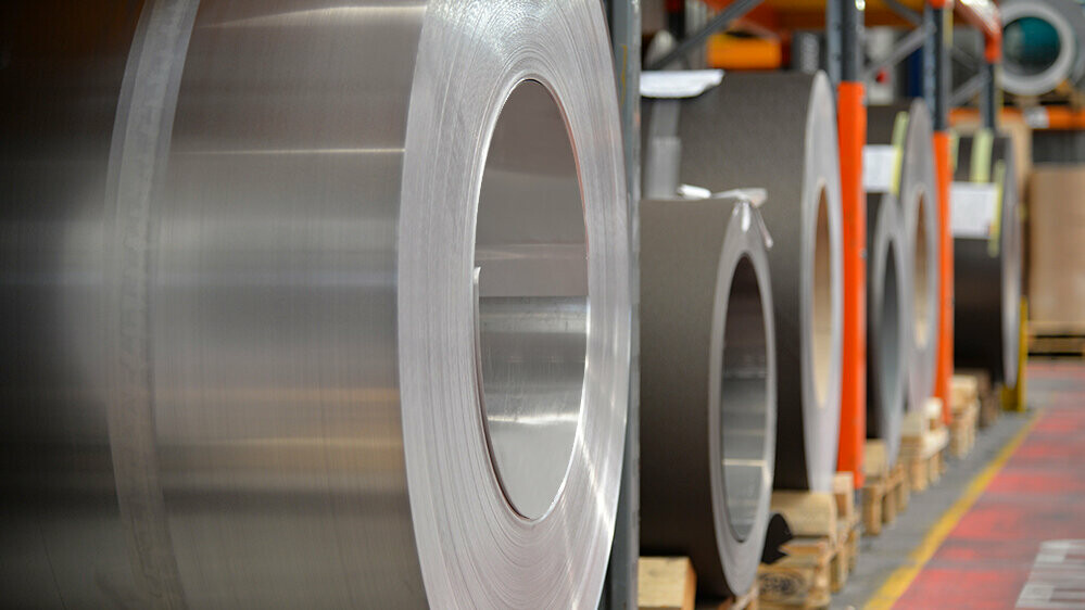 Aluminium rollen (coils) op houten palletten, op de voorgrond staat een aluminium rol in blank naturel, en daarachter zijn nog meer banden en platen van aluminium te zien.