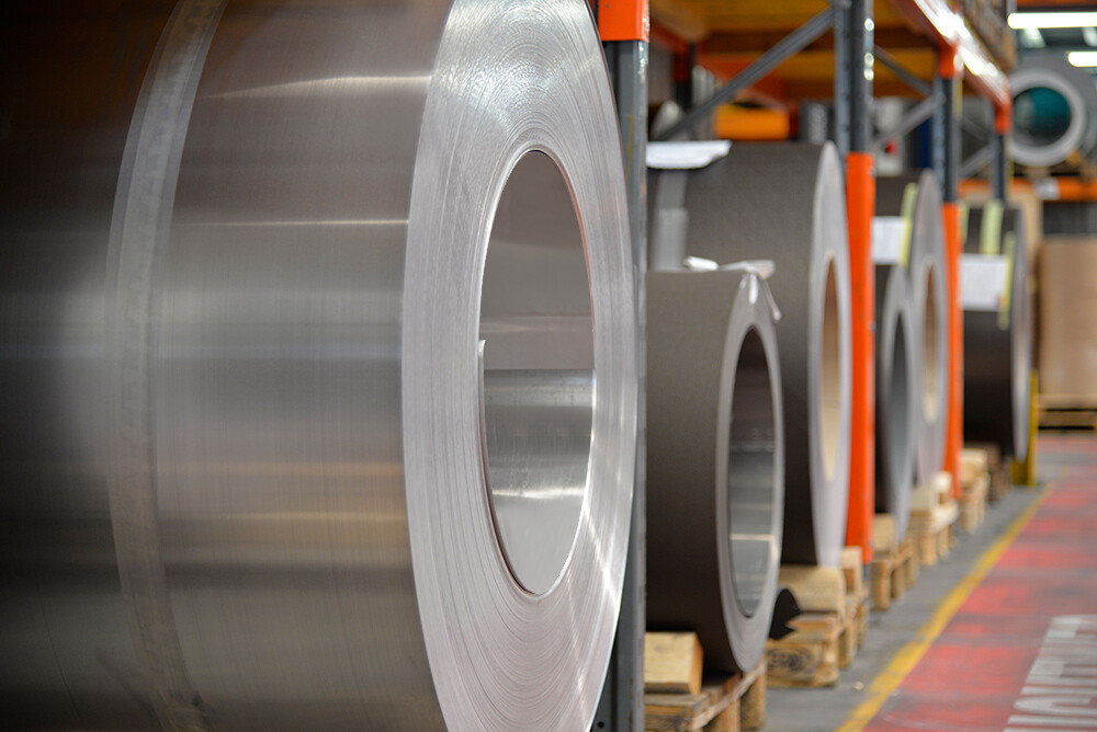 Aluminium rollen (coils) op houten palletten, op de voorgrond staat een aluminium rol in blank naturel, en daarachter zijn nog meer banden en platen van aluminium te zien.