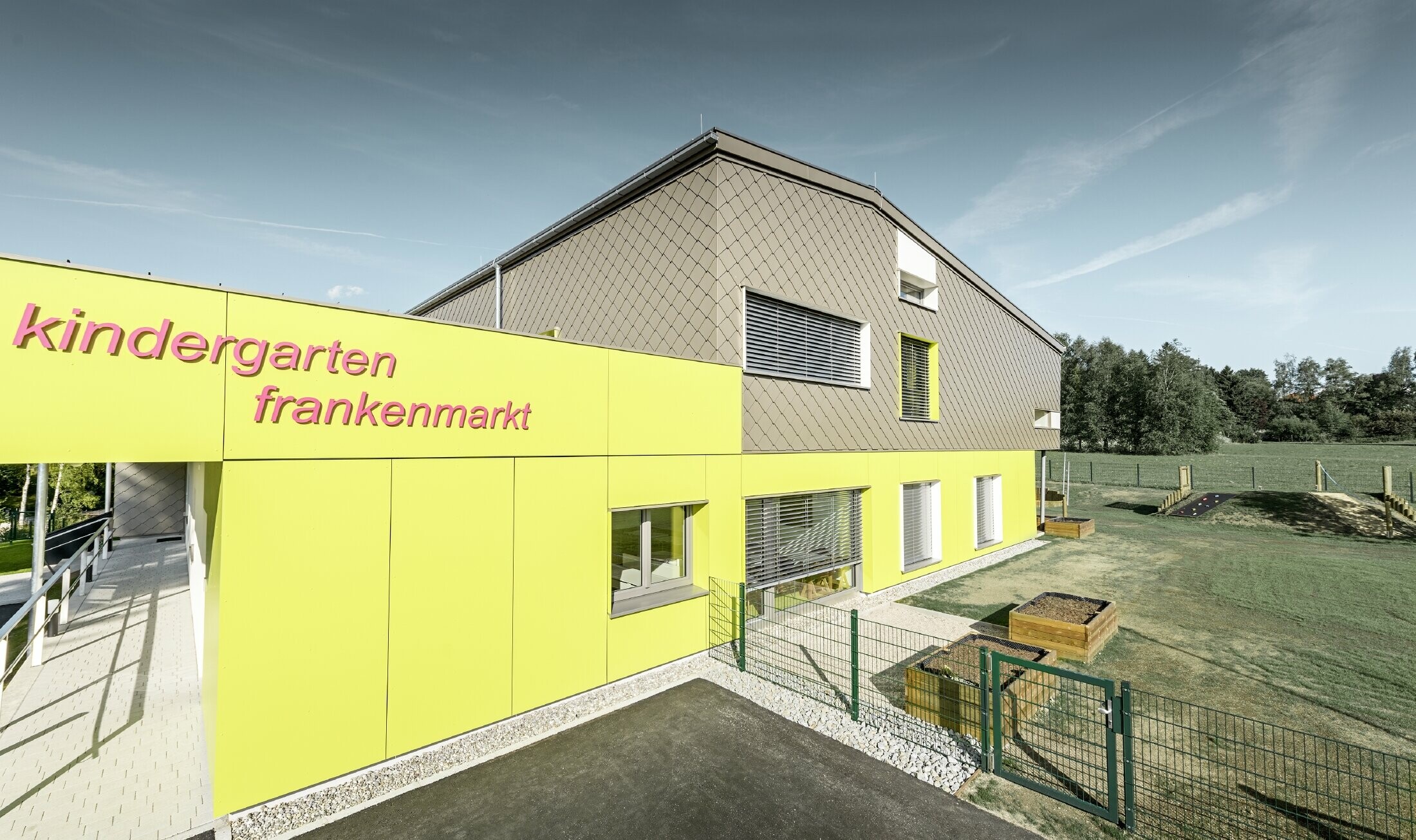 De gevel van de kleuterschool in Frankenmarkt is deels bekleed met de PREFA gevellosange in brons. De rest van de gevel is bekleed met gevelplaten in geel. De belettering van de kinderopvang is herkenbaar.