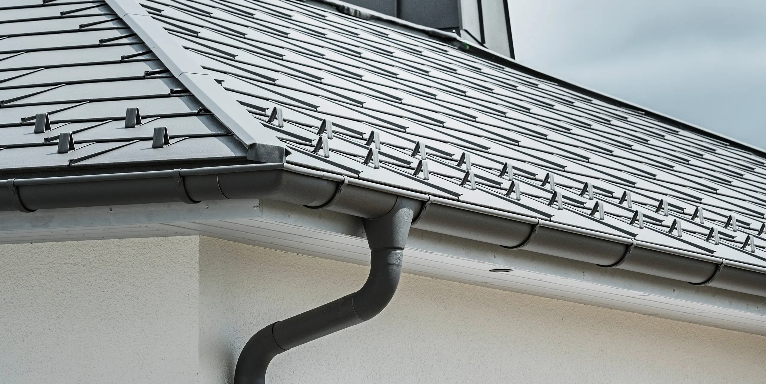 Close-up van de dakrand van een eengezinswoning, voorzien van het efficiënte PREFA dakafvoersysteem in P.10 donkergrijs. De afbeelding toont de gedetailleerd gelegde PREFA dakpan R.16 en daaronder de naadloos geïntegreerde mastgoot en afvoerbuis, die samen een betrouwbare en esthetisch aantrekkelijke oplossing bieden voor de afvoer van regenwater. De robuuste aluminium producten en de zorgvuldige montage onderstrepen de kwaliteit en de lange levensduur van het PREFA compleetsysteem.