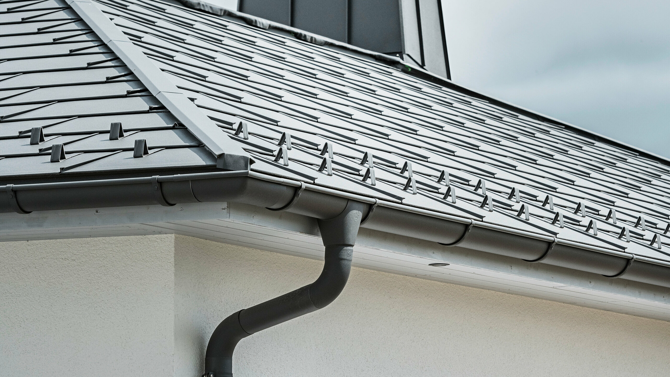 Close-up van de dakrand van een eengezinswoning, voorzien van het efficiënte PREFA dakafvoersysteem in P.10 donkergrijs. De afbeelding toont de gedetailleerd gelegde PREFA dakpan R.16 en daaronder de naadloos geïntegreerde mastgoot en afvoerbuis, die samen een betrouwbare en esthetisch aantrekkelijke oplossing bieden voor de afvoer van regenwater. De robuuste aluminium producten en de zorgvuldige montage onderstrepen de kwaliteit en de lange levensduur van het PREFA compleetsysteem.