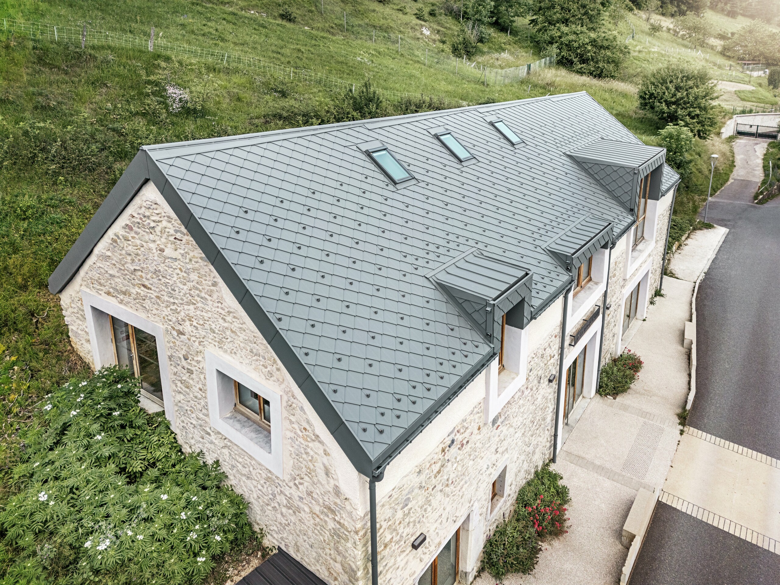 Dachansicht der restaurierten Scheune "La Grange à Gaby" in Engins, Frankreich. Das moderne PREFA Aluminiumdach mit Dachraute 29 × 29 fügt sich harmonisch in das historische Steingebäude ein und bietet langlebigen Schutz vor Witterungseinflüssen.
