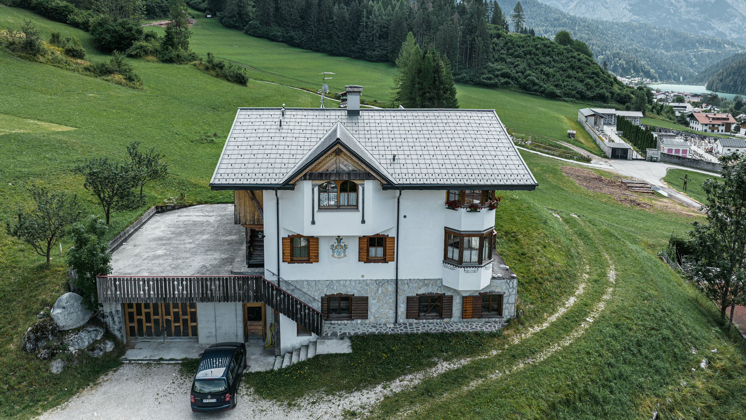 Vogelvluchtperspectief van de ingang van de traditionele berghut in Auronzo di Cadore na de dakrenovatie met PREFA aluminium dakpannen in P.10 lichtgrijs. Het gebouw met zijn karakteristieke alpine charme en het moderne tinnen dak past harmonisch in het weelderige groene landschap en het indrukwekkende bergpanorama op de achtergrond en toont een geslaagde combinatie van functionaliteit en regionale architectuur.