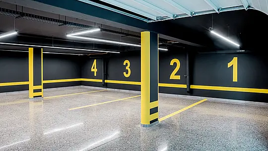 Die Aufnahme zeigt die moderne Tiefgarage der Budapester Mehrgenerationen‑Villa, gestaltet in einem klaren, industriellen Design mit dunklen Wänden und kontrastierenden gelben Markierungen. Die nummerierten Stellplätze und die präzise Linienführung vermitteln Struktur und Ordnung. Glänzender, polierter Boden reflektiert das Licht der schlanken LED‑Leuchten und betont die hochwertige Ausführung des unterirdischen Bereichs. Die Tiefgarage ergänzt das architektonisch anspruchsvolle Gebäude, dessen äußere Hülle durch langlebige, witterungsbeständige PREFA Aluminiumprodukte geprägt ist.