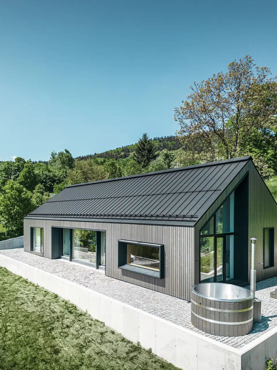 Ein modernes Holzmassivhaus in Kuncice (CZ) wurde mit großen Fensterflächen ausgestattet. Das dunkle Holzmassivhaus verfügt über ein Dachsystem aus PREFALZ in der Farbe P. 10 Schwarz. Im hinteren Außenbereich steht ein Whirlpool mit einer Holzverkleidung.