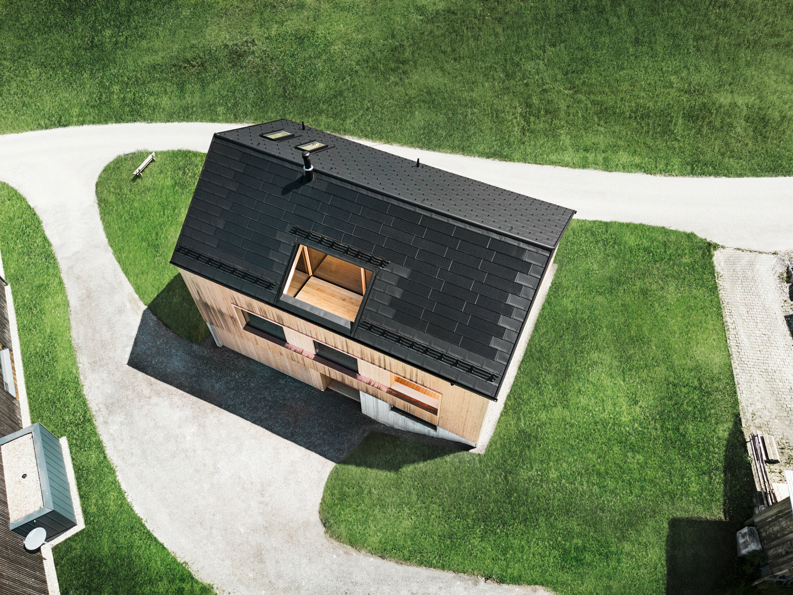 Drohnenaufnahme mit Blick von oben auf ein ökologisches Einfamilienhaus in Egg, das sich mit seinem schwarzen PREFA Solardach und den traditionellen PREFA Dachplatten R.16 von den umliegenden grünen Rasenflächen abhebt. Die strategisch angeordneten Solarmodule maximieren die Energieeffizienz, während die klare Geometrie des Daches und die natürlich wirkende Holzfassade eine Verbindung zwischen modernem Wohnen und der ländlichen Umgebung schaffen. Dieses Haus verkörpert eine fortschrittliche, umweltbewusste Bauweise, die sowohl funktional als auch optisch beeindruckt.