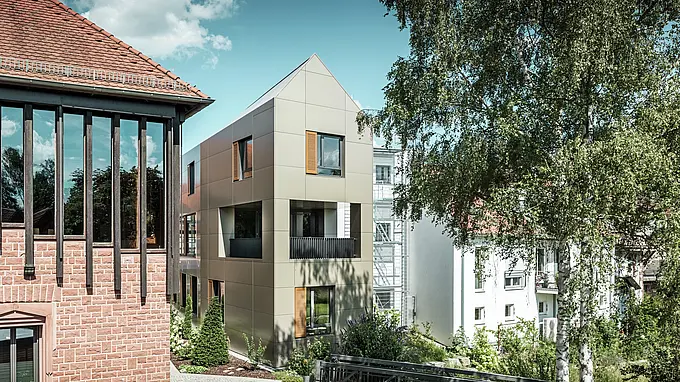 Translate to Dutch: Das Bild zeigt das moderne Rathaus in Alzenau, dessen Fassade aus Aluminium-Verbundplatten von PREFA in Bronze besteht. Der spitze Dachbereich und die klar strukturierten Fenster mit Holzjalousien verleihen dem Gebäude eine elegante und zeitgenössische Ästhetik. Im Vordergrund ist ein grüner Bereich mit einem gepflasterten Weg und Bäumen zu sehen, der das urbane Design des Gebäudes harmonisch ergänzt.