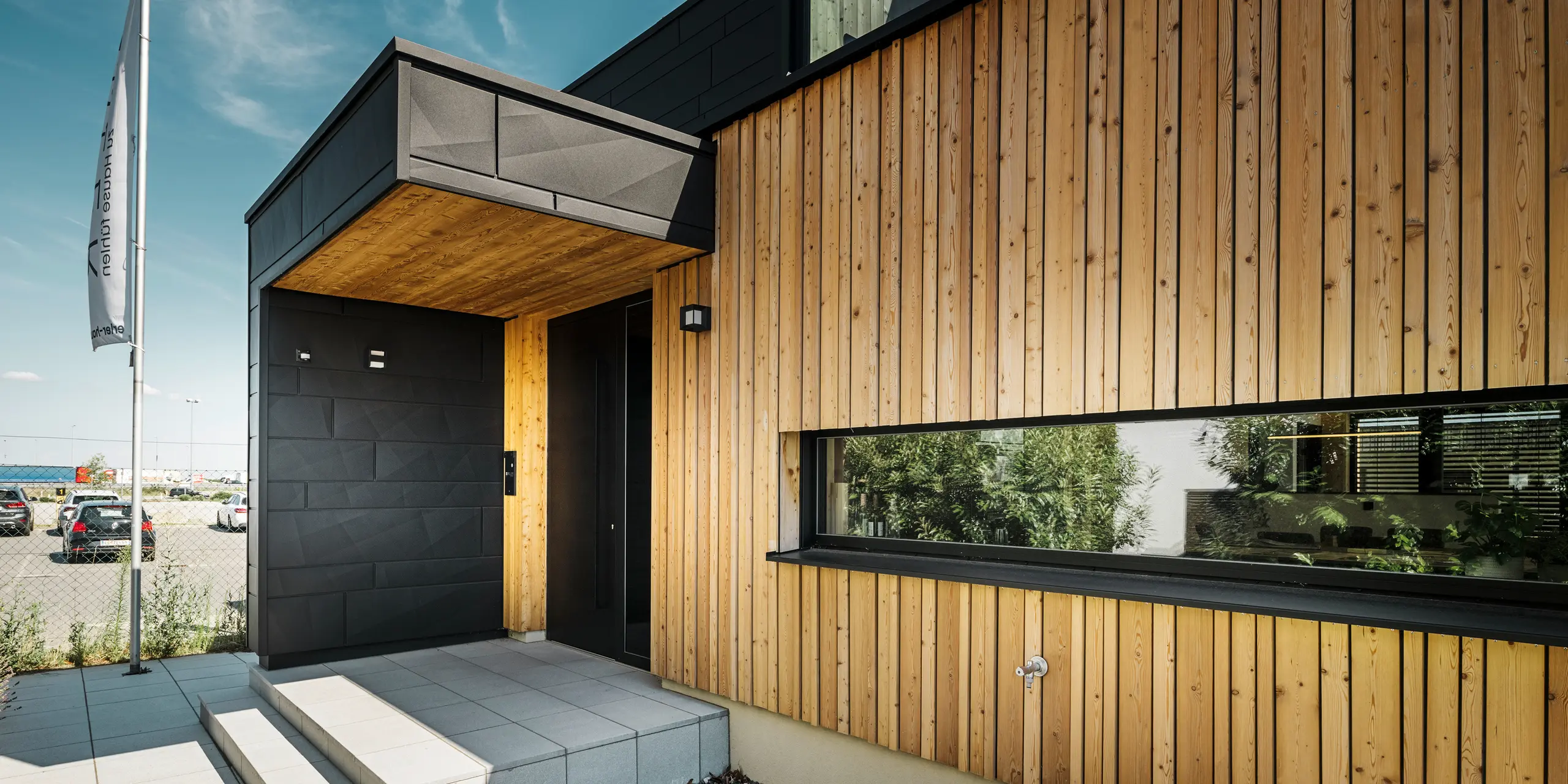 Zij-ingang van de Autark-showwoning in het prefabhuiscentrum Blaue Lagune in Vösendorf, met een moderne gevel van natuurlijk hout en zwarte PREFA Siding.X-platen. De foto toont de elegante combinatie van houten gevelbekleding en aluminium in de hoogwaardige coating P.10 Black.