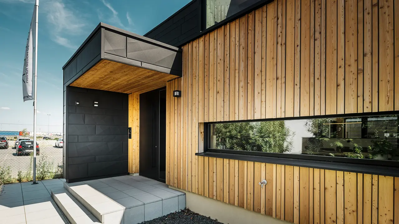 Zij-ingang van de Autark-showwoning in het prefabhuiscentrum Blaue Lagune in Vösendorf, met een moderne gevel van natuurlijk hout en zwarte PREFA Siding.X-platen. De foto toont de elegante combinatie van houten gevelbekleding en aluminium in de hoogwaardige coating P.10 Black.