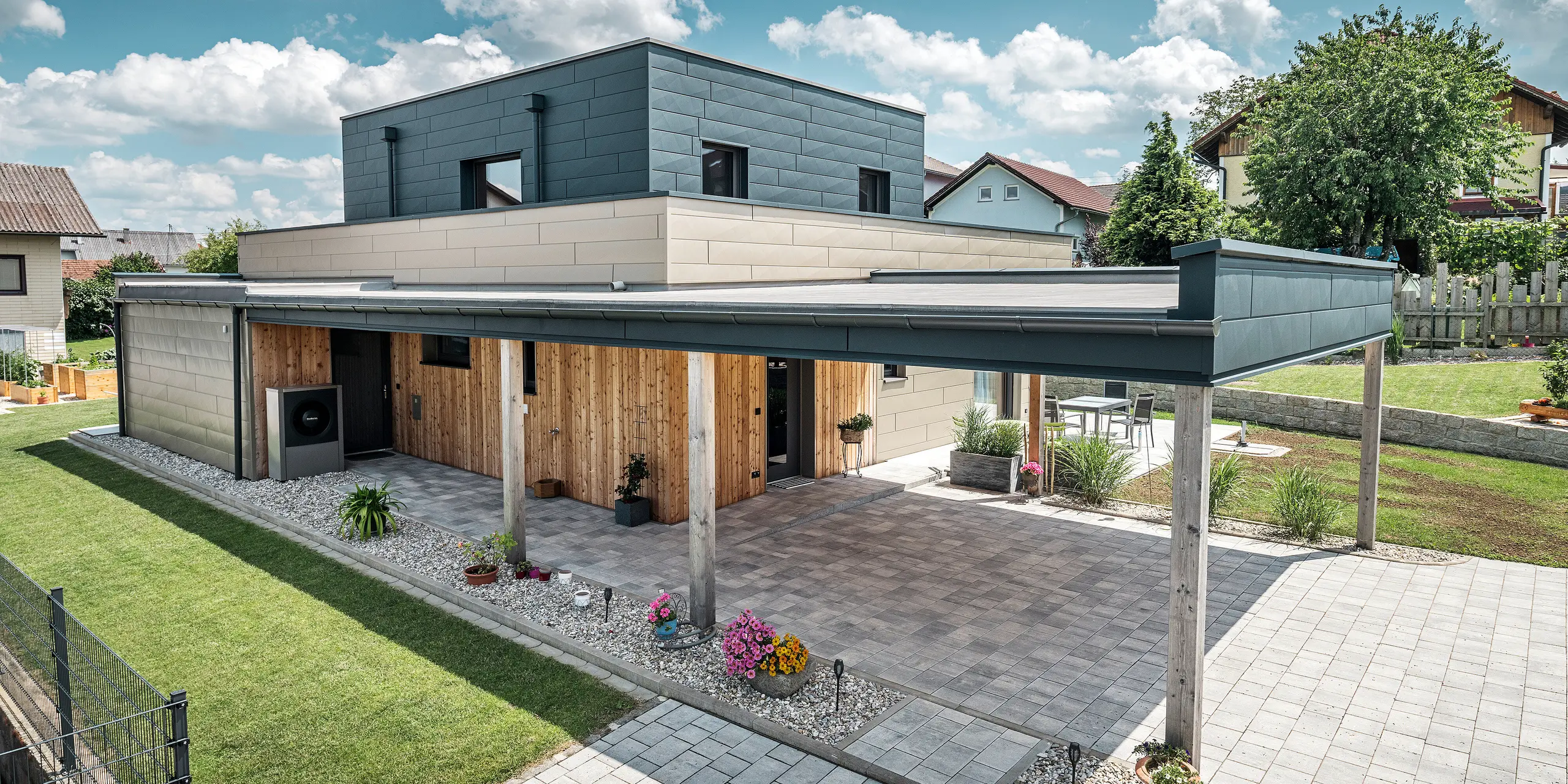 Luchtfoto van de oprit van een moderne eengezinswoning in Wippenham, Oostenrijk, met een tweekleurige PREFA aluminium gevel in brons en antraciet. De woning toont de stijlvolle integratie van Siding.X gevelpaneelen en houten bekleding. Duidelijk zichtbaar is de overdekte carport, die naadloos is geïntegreerd in de architectuur van het huis. De verzorgde tuin en het terras onderstrepen het moderne en functionele ontwerp van het gebouw.