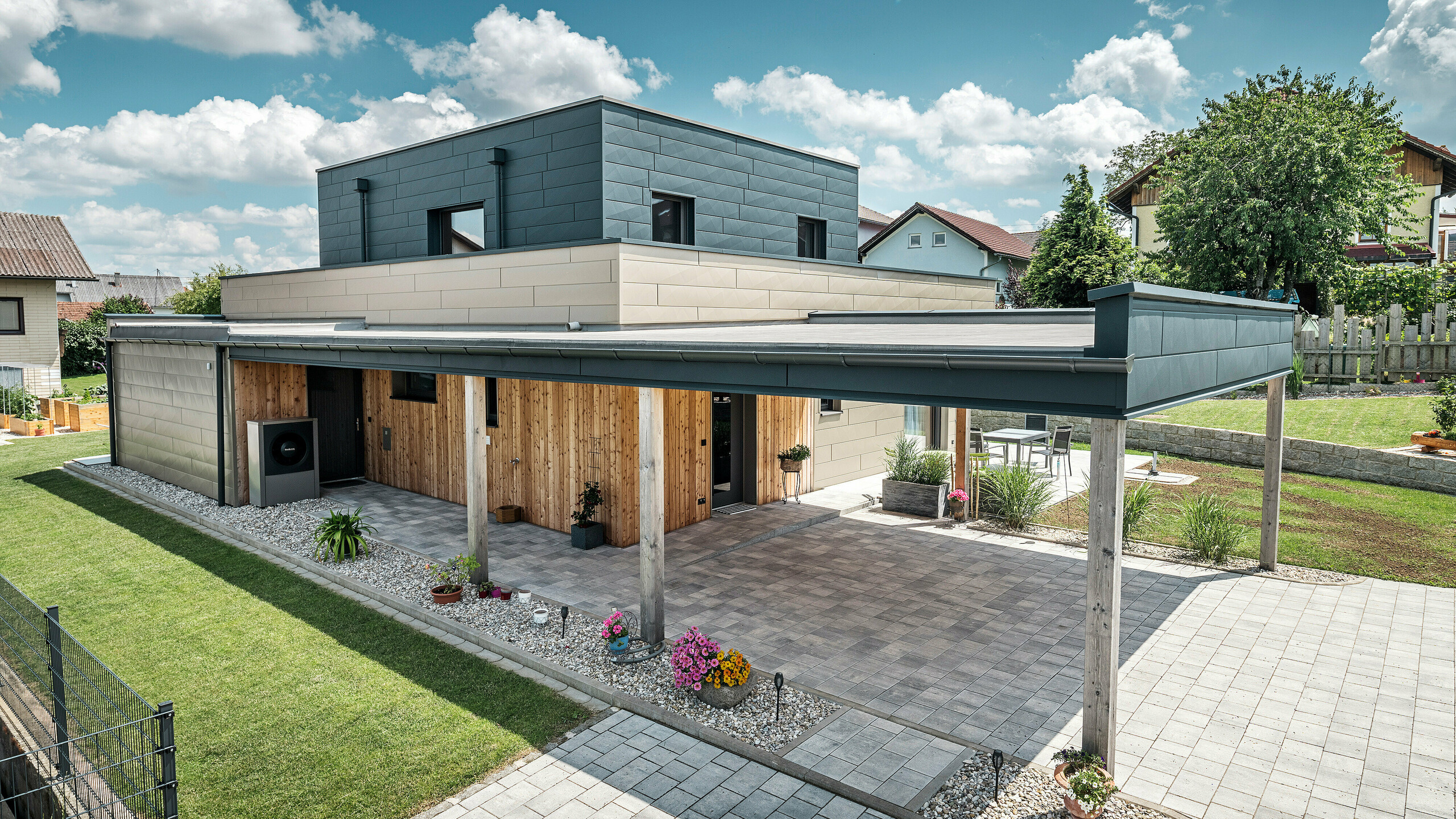 Luchtfoto van de oprit van een moderne eengezinswoning in Wippenham, Oostenrijk, met een tweekleurige PREFA aluminium gevel in brons en antraciet. De woning toont de stijlvolle integratie van Siding.X gevelpaneelen en houten bekleding. Duidelijk zichtbaar is de overdekte carport, die naadloos is geïntegreerd in de architectuur van het huis. De verzorgde tuin en het terras onderstrepen het moderne en functionele ontwerp van het gebouw.
