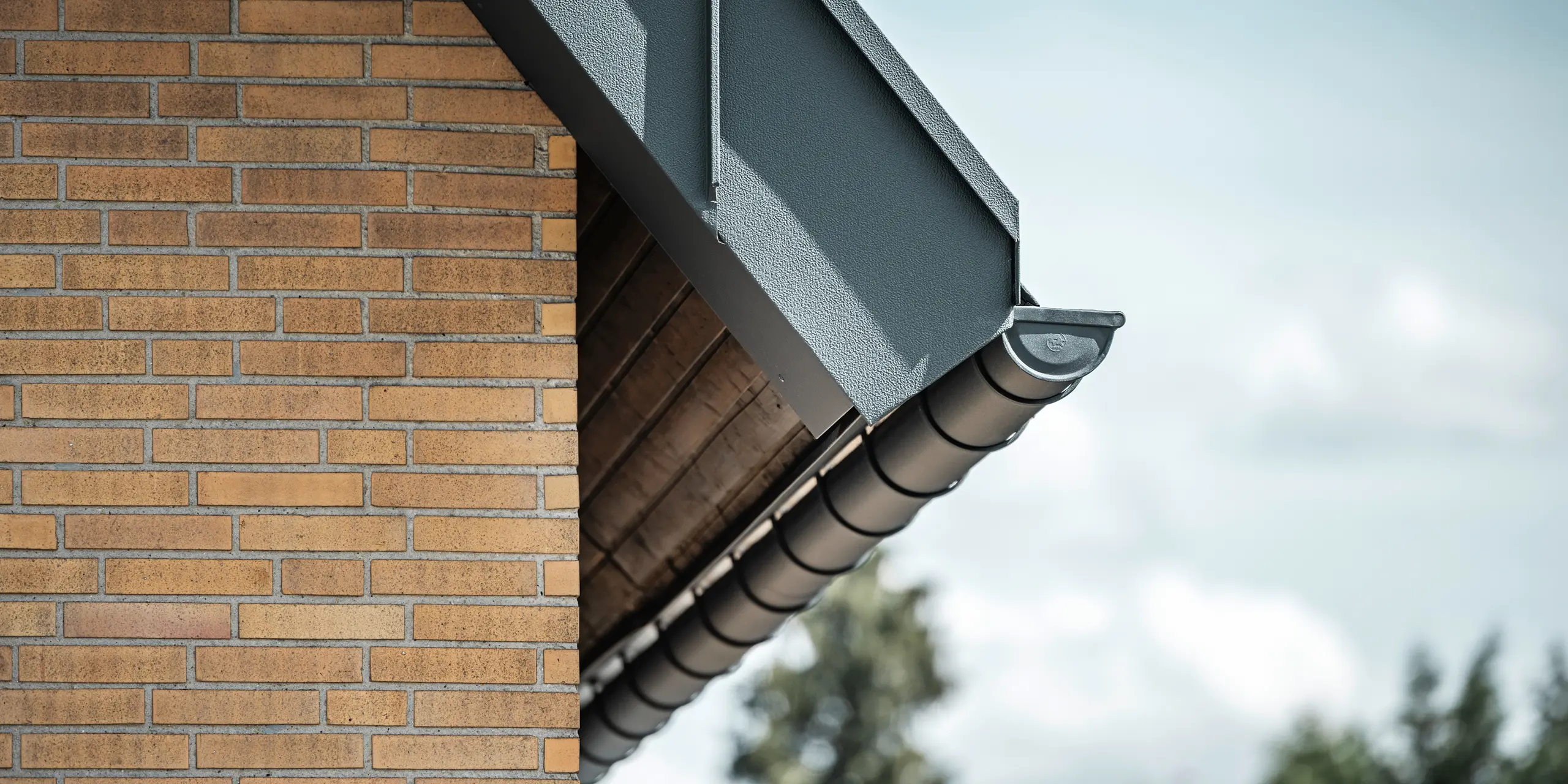 Detailopname van een antracietkleurig dakafvoersysteem van PREFA. De mastgoot steekt duidelijk af tegen de warme tinten van de bakstenen muur en benadrukt de nauwkeurige afwerking en kwaliteit van de gebruikte materialen.