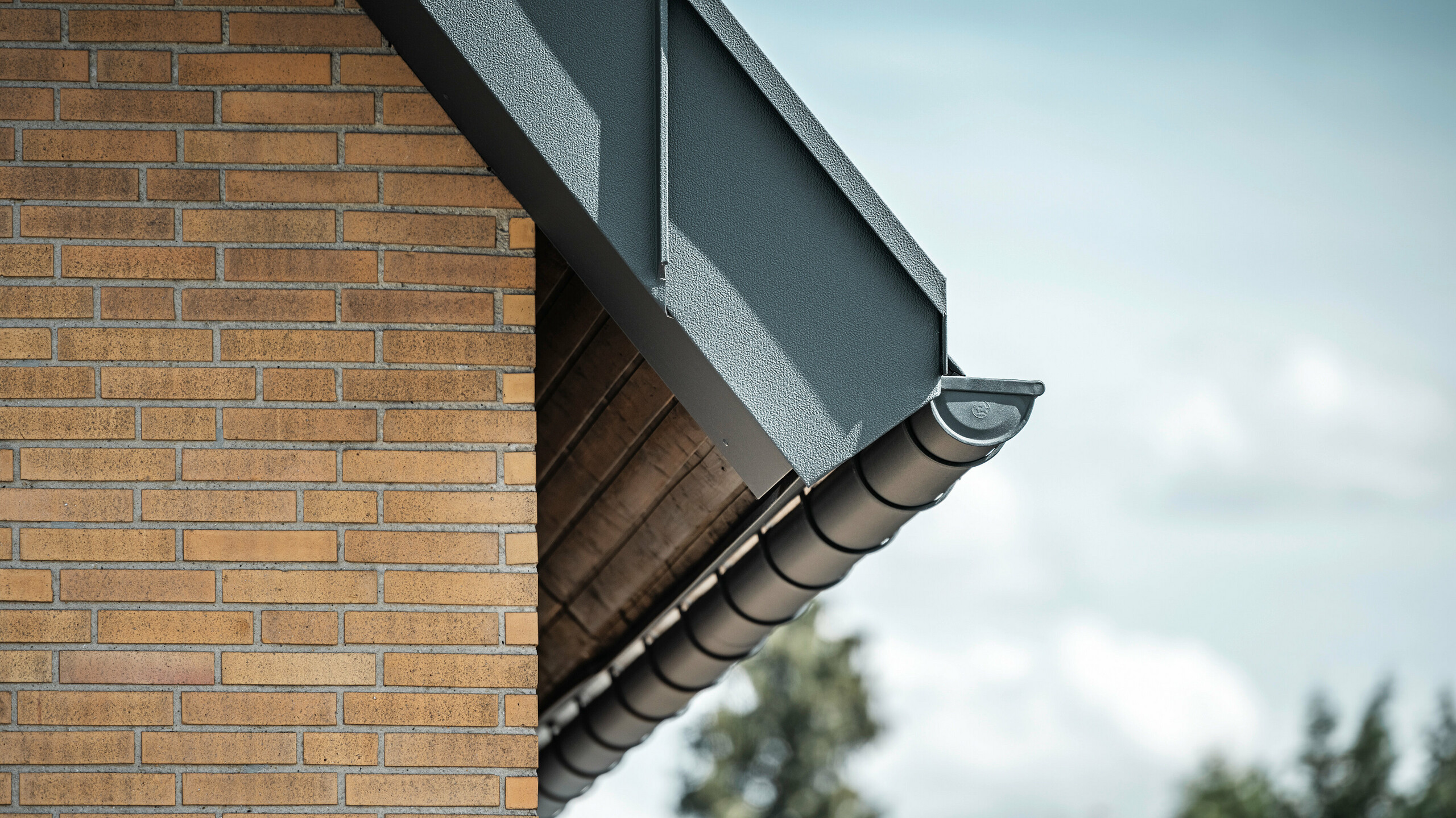 Detailopname van een antracietkleurig dakafvoersysteem van PREFA. De mastgoot steekt duidelijk af tegen de warme tinten van de bakstenen muur en benadrukt de nauwkeurige afwerking en kwaliteit van de gebruikte materialen.