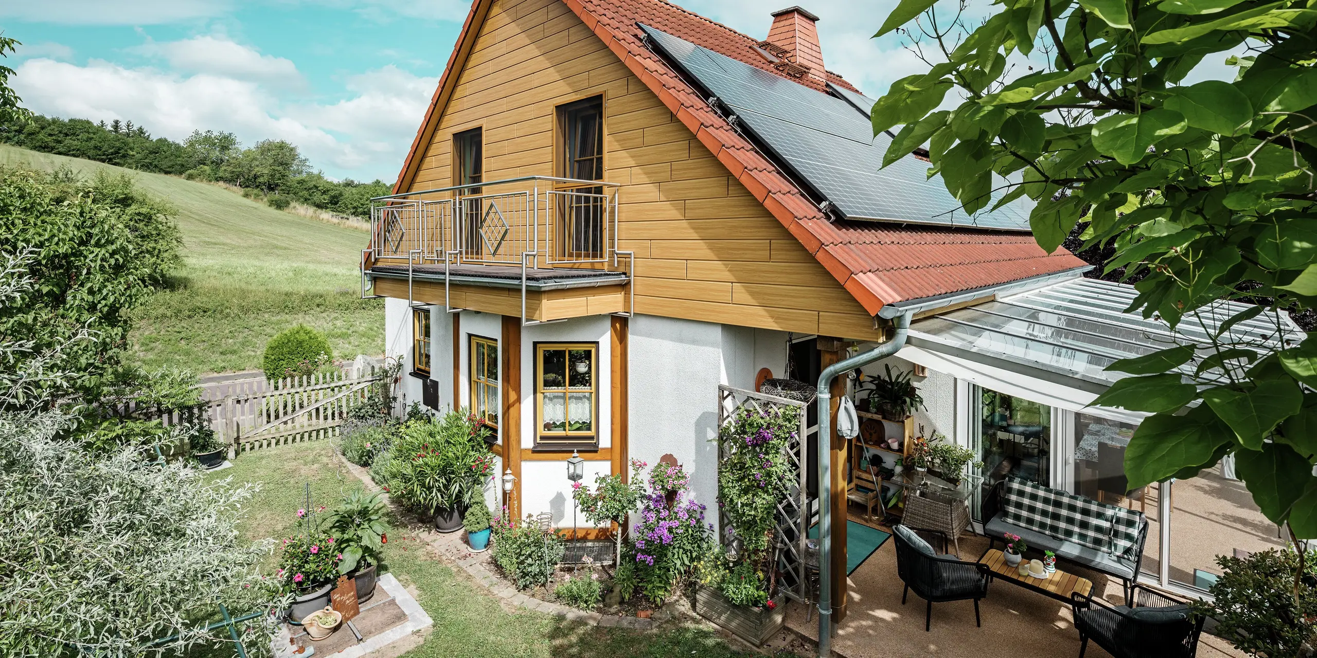 Deze afbeelding toont een charmante eengezinswoning in Blieskastel, met een pas gerenoveerde puntgevel met PREFA Sidings in eiken naturel die onmiddellijk in het oog springt. De warme houtlook van duurzaam aluminium sluit indrukwekkend mooi aan op het rode pannendak en de grote veranda. Te midden van weelderig groen en liefdevol aangelegde bloemperken ontstaat een idyllisch, levendig tafereel. De robuuste gevelrenovatie met PREFA geeft het huis niet gewoon een natuurlijke look, maar overtuigt ook door weerbestendigheid, kleurechtheid en een nagenoeg onderhoudsvrije en duurzame kwaliteit.