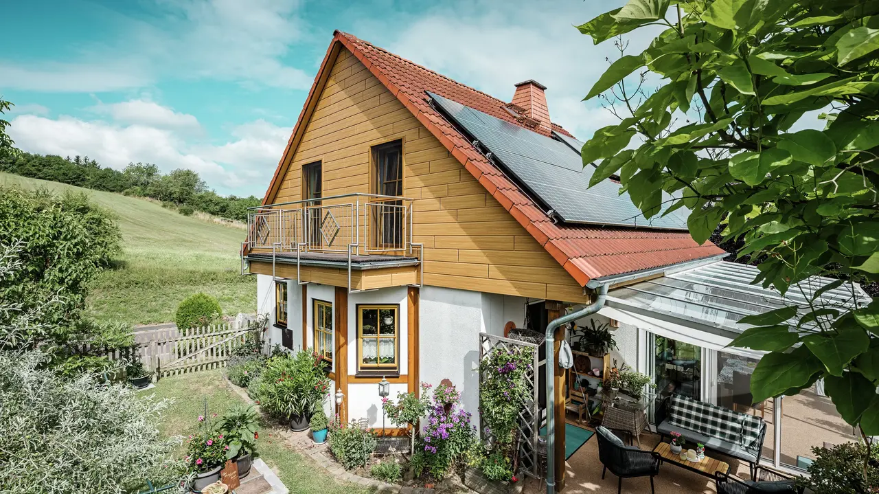 Deze afbeelding toont een charmante eengezinswoning in Blieskastel, met een pas gerenoveerde puntgevel met PREFA Sidings in eiken naturel die onmiddellijk in het oog springt. De warme houtlook van duurzaam aluminium sluit indrukwekkend mooi aan op het rode pannendak en de grote veranda. Te midden van weelderig groen en liefdevol aangelegde bloemperken ontstaat een idyllisch, levendig tafereel. De robuuste gevelrenovatie met PREFA geeft het huis niet gewoon een natuurlijke look, maar overtuigt ook door weerbestendigheid, kleurechtheid en een nagenoeg onderhoudsvrije en duurzame kwaliteit.