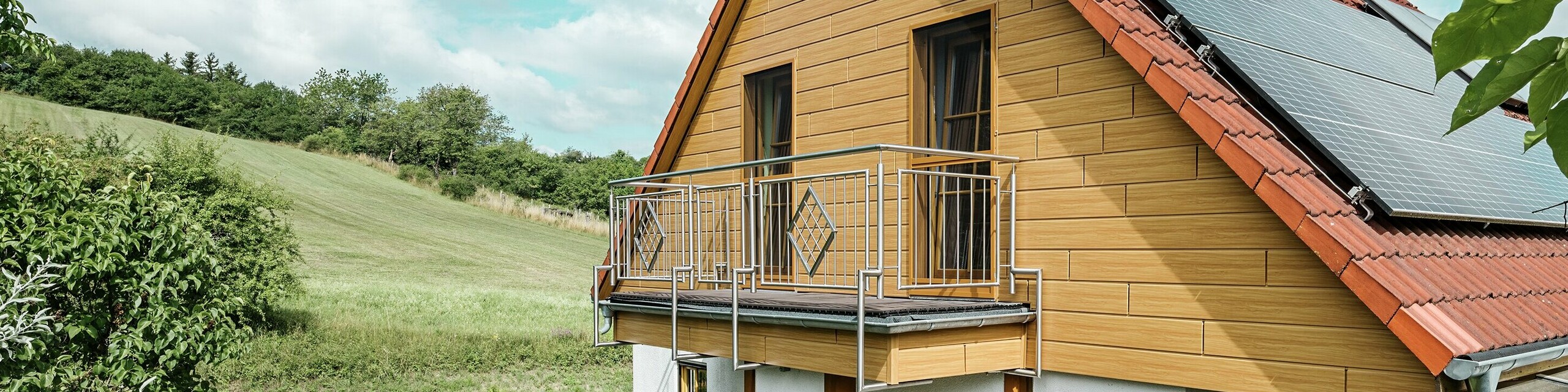 Deze afbeelding toont een charmante eengezinswoning in Blieskastel, met een pas gerenoveerde puntgevel met PREFA Sidings in eiken naturel die onmiddellijk in het oog springt. De warme houtlook van duurzaam aluminium sluit indrukwekkend mooi aan op het rode pannendak en de grote veranda. Te midden van weelderig groen en liefdevol aangelegde bloemperken ontstaat een idyllisch, levendig tafereel. De robuuste gevelrenovatie met PREFA geeft het huis niet gewoon een natuurlijke look, maar overtuigt ook door weerbestendigheid, kleurechtheid en een nagenoeg onderhoudsvrije en duurzame kwaliteit.