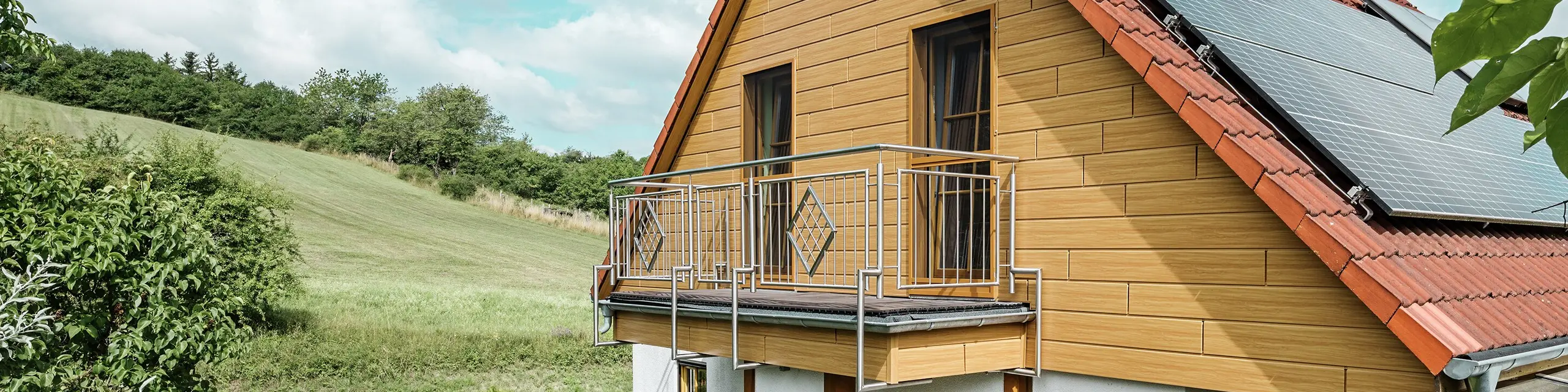 Deze afbeelding toont een charmante eengezinswoning in Blieskastel, met een pas gerenoveerde puntgevel met PREFA Sidings in eiken naturel die onmiddellijk in het oog springt. De warme houtlook van duurzaam aluminium sluit indrukwekkend mooi aan op het rode pannendak en de grote veranda. Te midden van weelderig groen en liefdevol aangelegde bloemperken ontstaat een idyllisch, levendig tafereel. De robuuste gevelrenovatie met PREFA geeft het huis niet gewoon een natuurlijke look, maar overtuigt ook door weerbestendigheid, kleurechtheid en een nagenoeg onderhoudsvrije en duurzame kwaliteit.