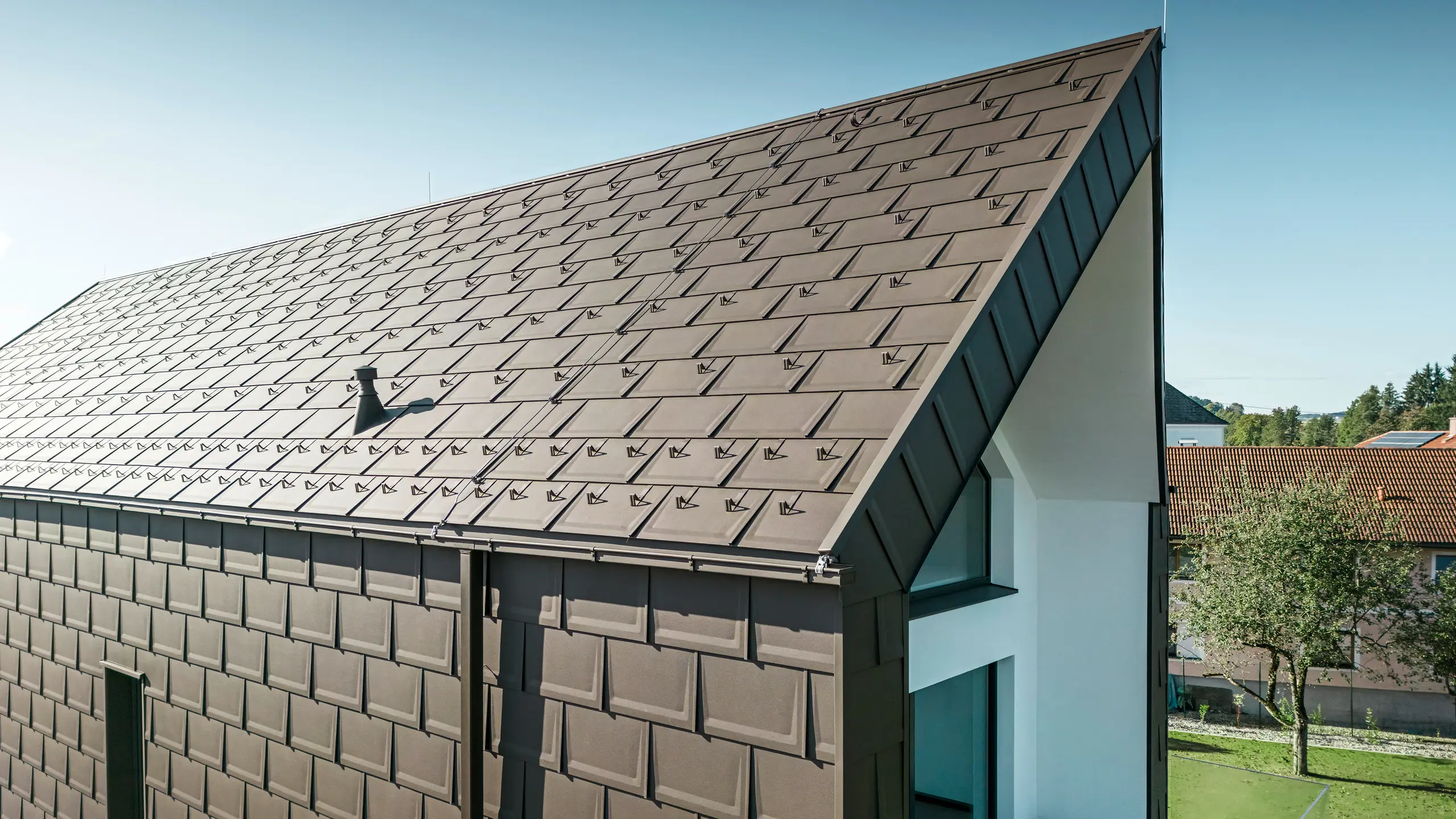 Een perspectivisch aanzicht van het dak en de gevel van een moderne eengezinswoning in Neukirchen, Oostenrijk. De PREFA R.16 dakpan in bruintint P.10 biedt een esthetische en functionele dakbedekking die perfect past bij het architectonische ontwerp van de woning. De scherpe nok van het dak en de nauwkeurige plaatsing van de dakpanelen tonen de kwaliteit van PREFA. De kleine formaten aluminium producten passen perfect bij de wit gepleisterde gevel en het omringende groene landschap.