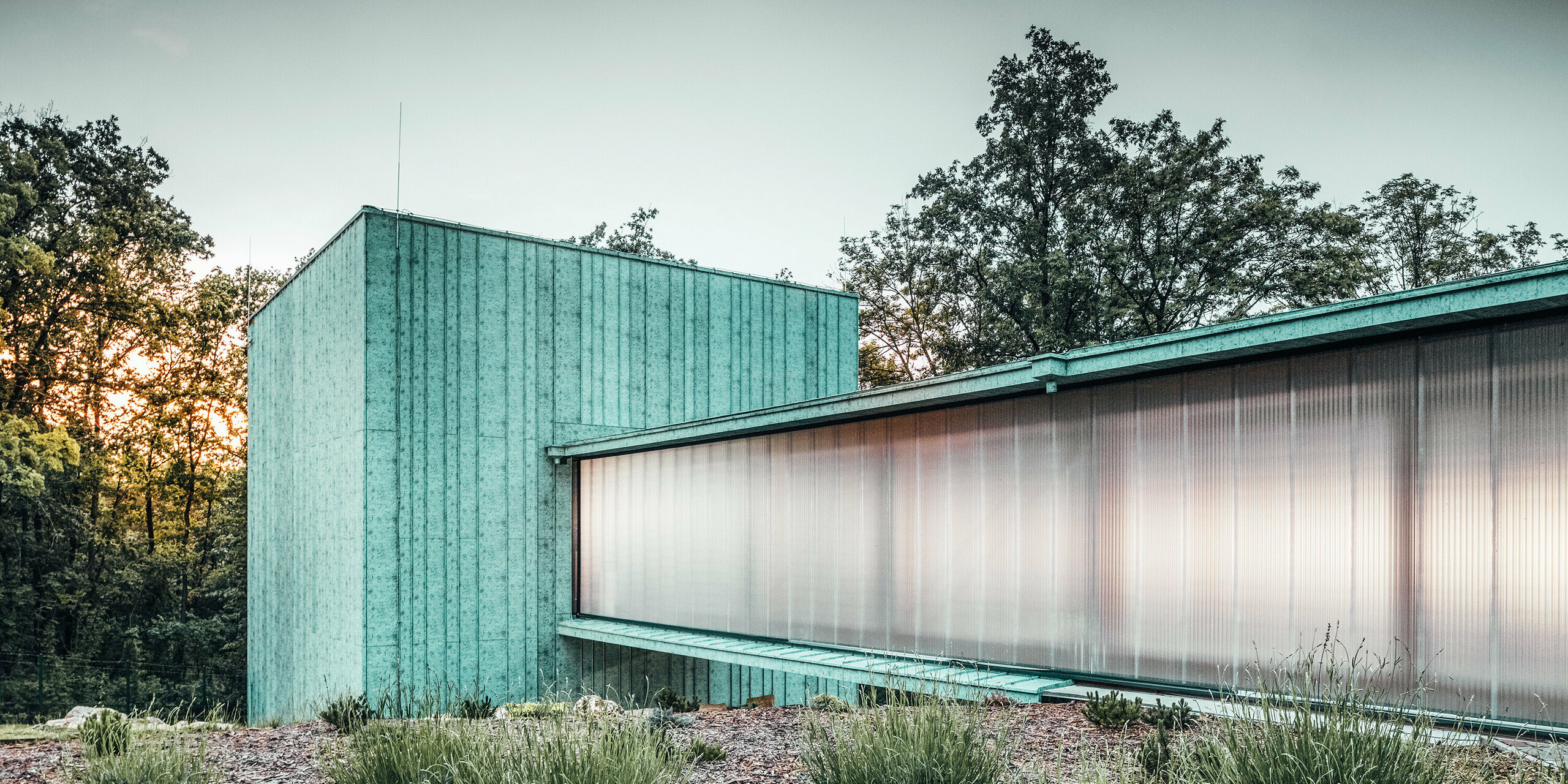 Das Zero Magnetic Field Laboratory in Fertőboz (HU) ist umhüllt von PREEFALZ in der Farbe P.10 Patinagrün. Das würfelförmige Laborgebäude durch einen Tunnel mit dem Nebengebäude verbunden. Die Seitenwände des Durchgangs verleihen dem Bauwerk eine Transparenz durch ihre Milchglasfolierung. Im Hintergrund ist die natürliche Landschaft im Licht des Sonnenuntergangs zu sehen. 