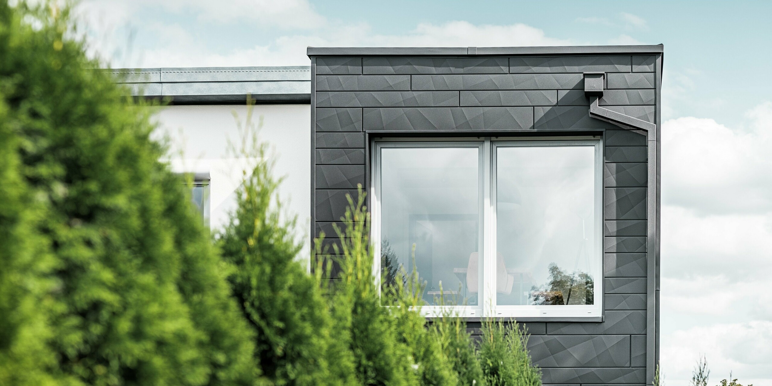 De duidelijk gestructureerde PREFA Siding.X gevelpaneelen in de kleur P.10 zwart sieren de buitenmuur van een eengezinswoning in Teuschnitz en geven deze een unieke uitstraling. De PREFA dakafvoersystemen, bestaande uit de vierkante buis en de vergaarbak, sluit naadloos aan op het ontwerp. De groene thuja's op de voorgrond contrasteren met de donkere metalen gevel.