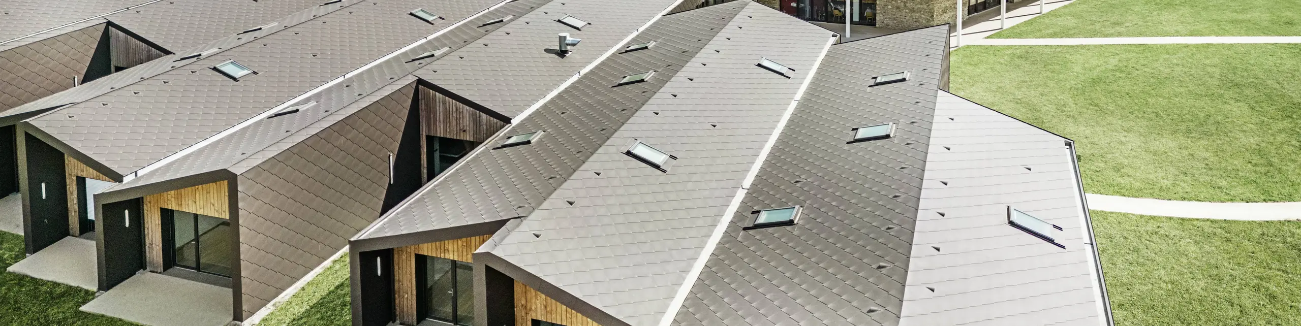 Das Bild zeigt eine Luftaufnahme einer modernen Wohnanlage mit Dach- und Wandelementen aus PREFA 44 × 44 in Walnussbraun. Die Dachlandschaft zeichnet sich durch geometrische Formen aus, die mit großen Glasflächen und Holzelementen kombiniert werden. Im Hintergrund ist eine alte Kirche zu sehen, die einen harmonischen Kontrast zur modernen Architektur bildet. Der grüne Rasen und die umliegende Natur unterstreichen das offene und nachhaltige Design der gesamten Anlage.