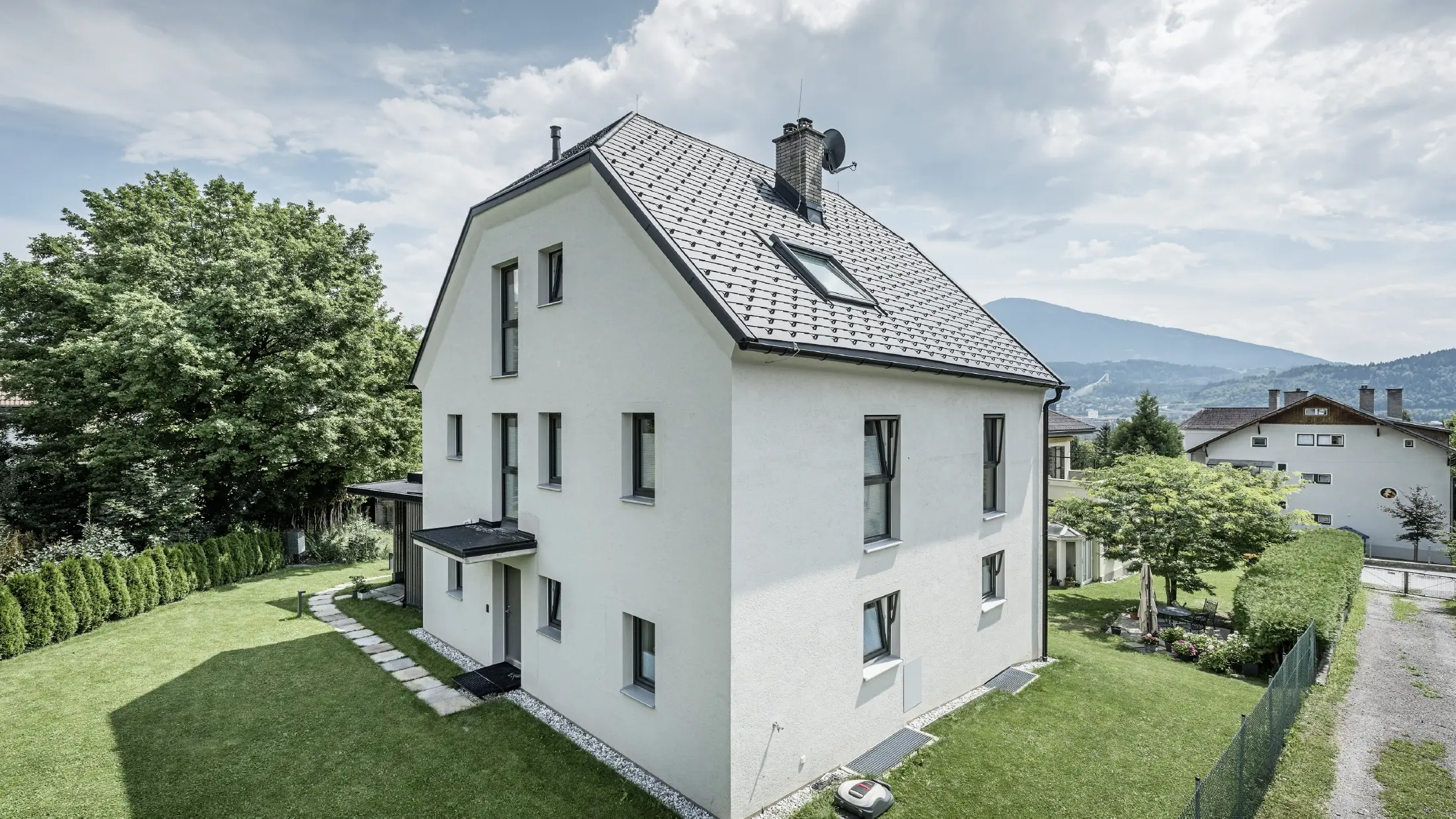 Moderne vrijstaande woning met drie verdiepingen, een lichte pleistergevel en een donkergrijs zadeldak van aluminium van PREFA. Het gebouw heeft meerdere smalle ramen en dakafvoersystemen in antraciet. Het staat in een verzorgde tuin met gazon, bomen en heggen. Op de achtergrond zijn nog meer woonhuizen en een bergachtig landschap te zien.
