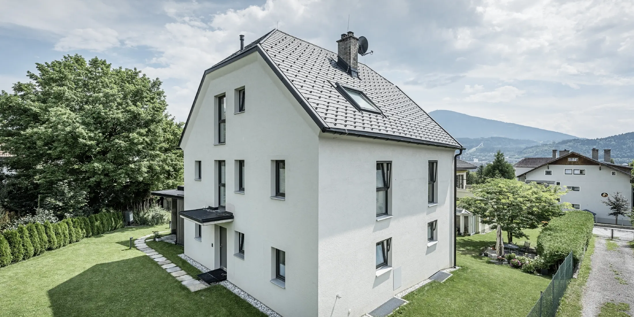 Moderne vrijstaande woning met drie verdiepingen, een lichte pleistergevel en een donkergrijs zadeldak van aluminium van PREFA. Het gebouw heeft meerdere smalle ramen en dakafvoersystemen in antraciet. Het staat in een verzorgde tuin met gazon, bomen en heggen. Op de achtergrond zijn nog meer woonhuizen en een bergachtig landschap te zien.