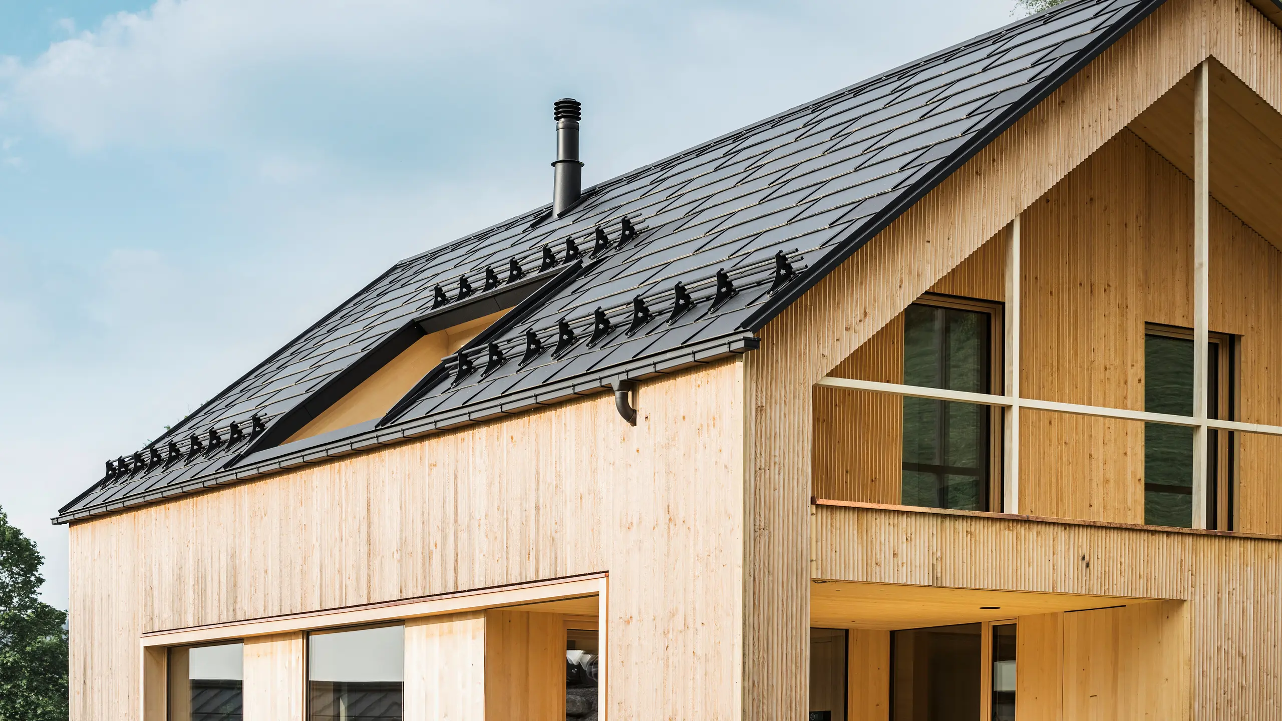 Close-up van de gevel van een milieuvriendelijke woning in Egg, gekenmerkt door PREFA solar-dakpannen in elegant zwart, die harmonieus zijn geïntegreerd in het midden van de PREFA dakpan R.16. De strakke lijnen van het dak contrasteren met de natuurlijke textuur van de houten gevel, terwijl de solarmodules een statement maken voor duurzaam wonen. De subtiele schoorsteen past in het totaalbeeld, dat milieubewust design combineert met landelijke esthetiek. Bovendien beschikt de innovatieve eco-woning over twee balkons die uitzicht bieden op het prachtige berglandschap van Egg in Vorarlberg.