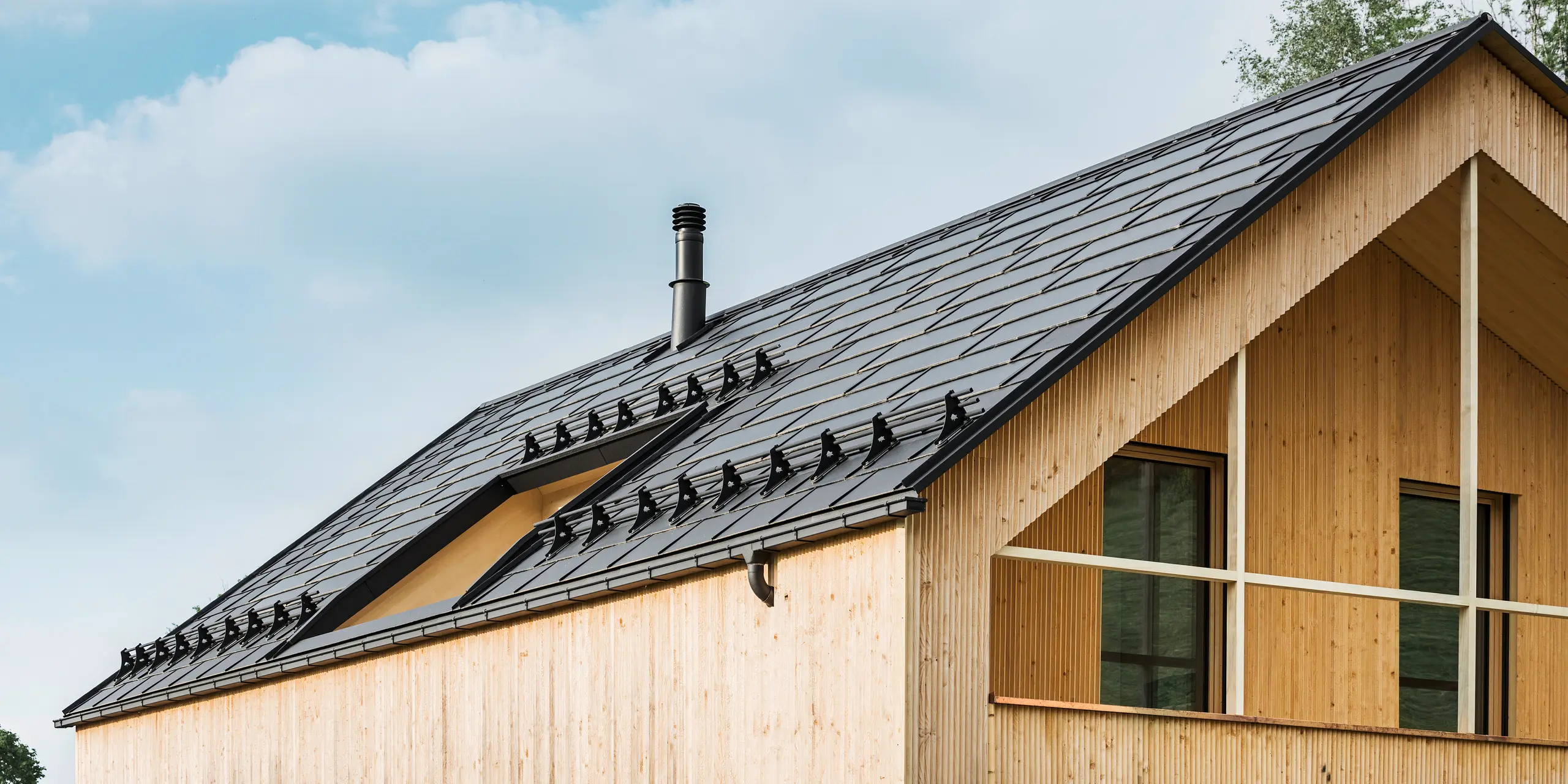 Close-up van de gevel van een milieuvriendelijke woning in Egg, gekenmerkt door PREFA solar-dakpannen in elegant zwart, die harmonieus zijn geïntegreerd in het midden van de PREFA dakpan R.16. De strakke lijnen van het dak contrasteren met de natuurlijke textuur van de houten gevel, terwijl de solarmodules een statement maken voor duurzaam wonen. De subtiele schoorsteen past in het totaalbeeld, dat milieubewust design combineert met landelijke esthetiek. Bovendien beschikt de innovatieve eco-woning over twee balkons die uitzicht bieden op het prachtige berglandschap van Egg in Vorarlberg.