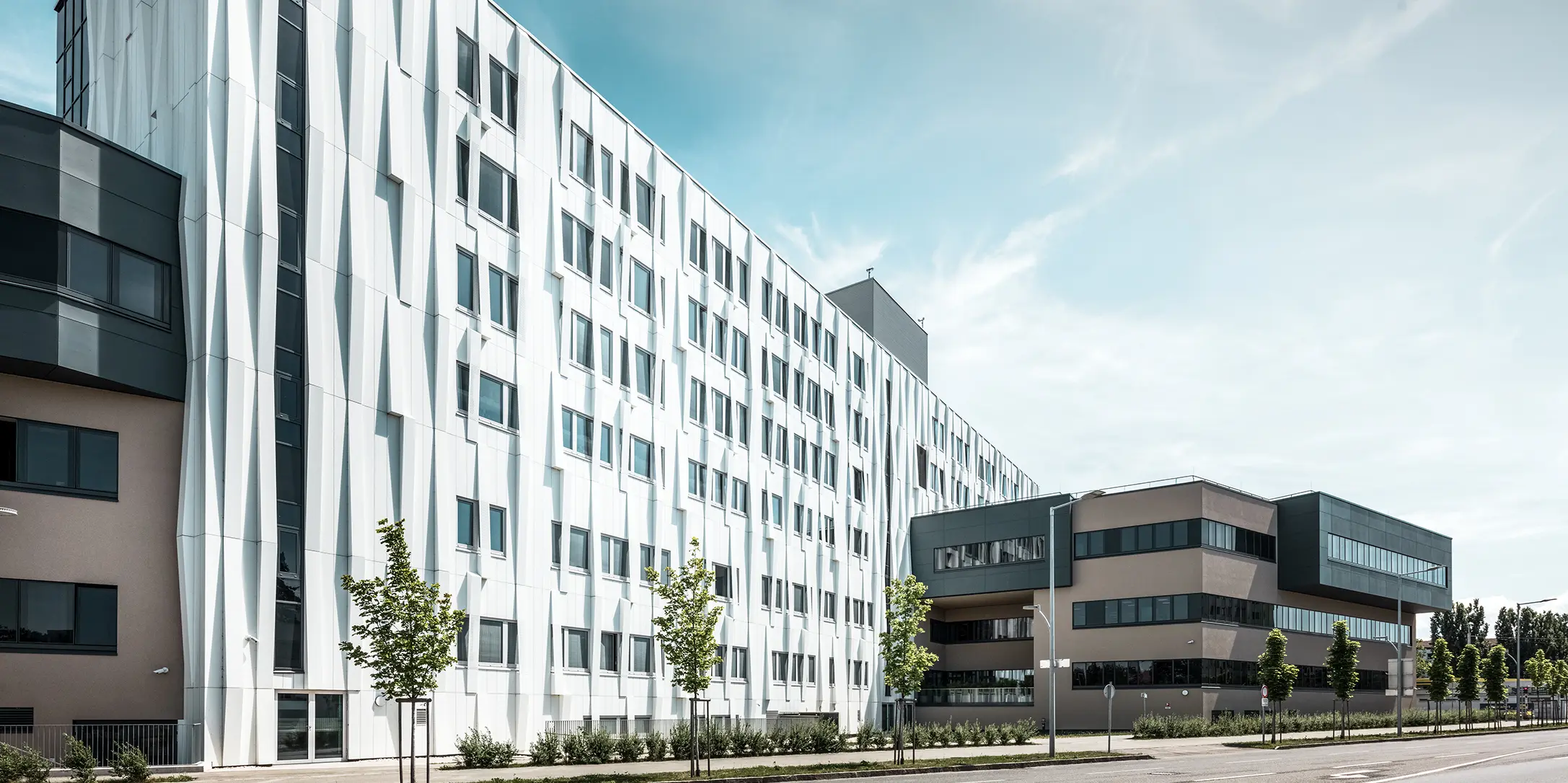 Zijaanzicht van het universitair ziekenhuis in St. Pölten, Neder-Oostenrijk. De robuuste aluminium gevel maakt het gebouw tot een blikvanger in de stad. Op de voorgrond is een straat met enkele bomen te zien.