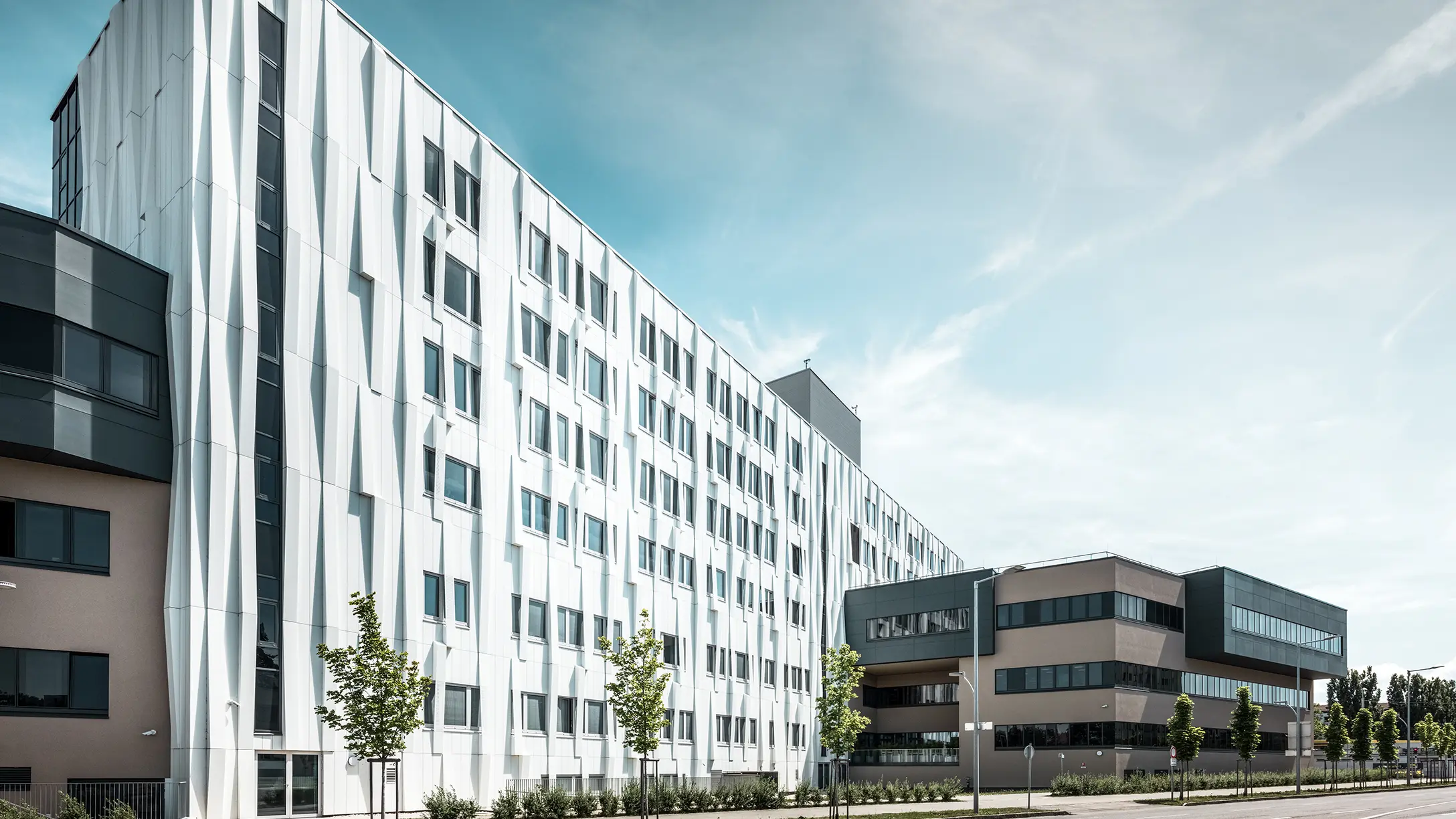 Zijaanzicht van het universitair ziekenhuis in St. Pölten, Neder-Oostenrijk. De robuuste aluminium gevel maakt het gebouw tot een blikvanger in de stad. Op de voorgrond is een straat met enkele bomen te zien.