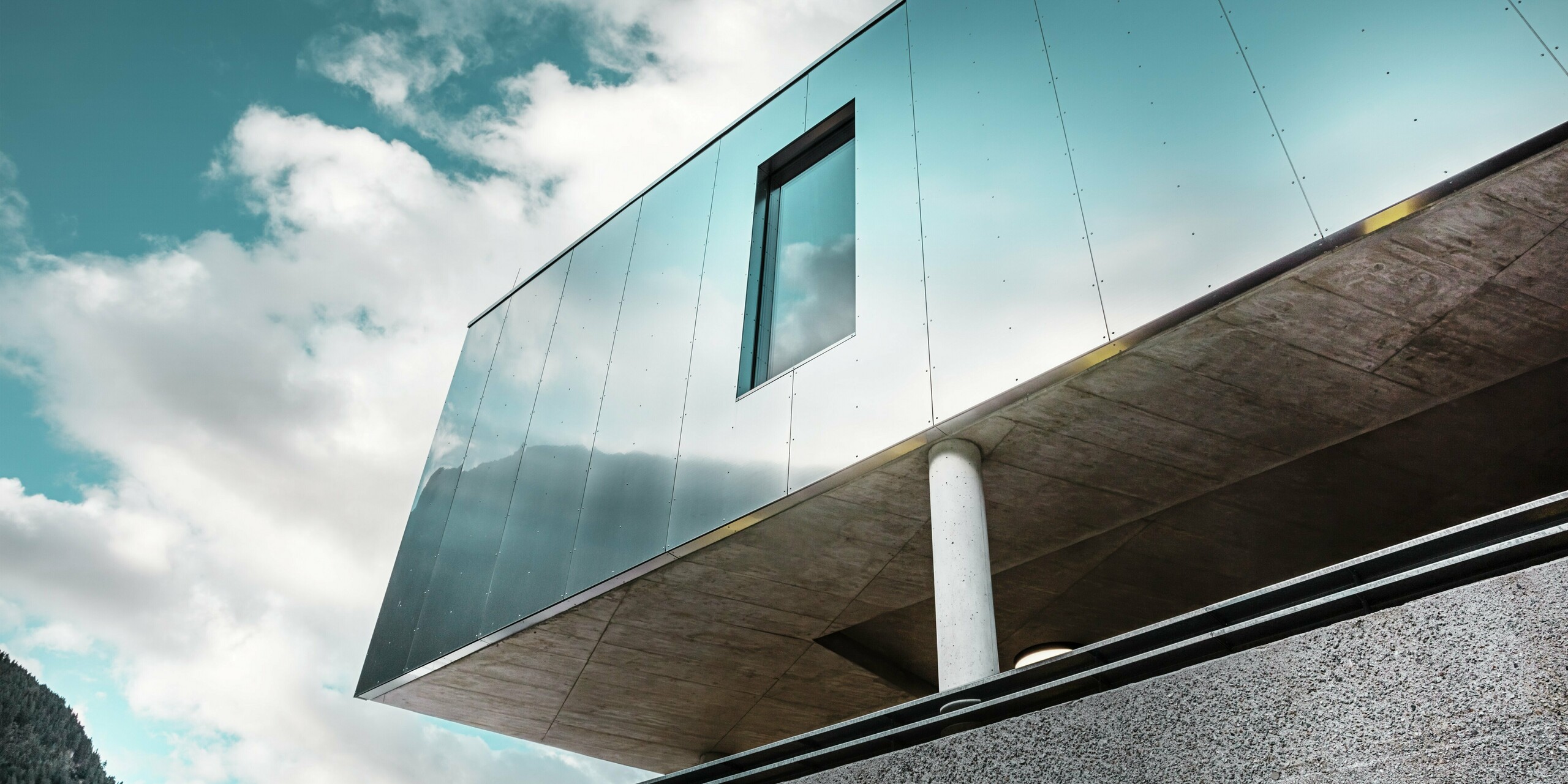 Moderne Architektur des MPREIS Supermarktes in Sölden, Tirol, mit reflektierenden PREFA Aluminium Verbundplatten in der Sonderfarbe Reflective Natural. Die glänzende Fassade spiegelt den Himmel und die Berglandschaft wider. Klare Linien und massive Betonsäulen verbinden innovatives Design mit Stabilität und Eleganz.
