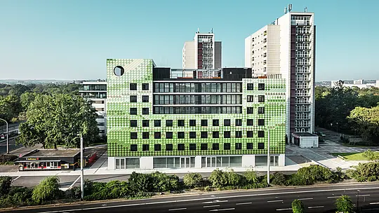 Seitenansicht des Wohn- und Geschäftshauses „Pick-Nick“ am Straßburger Platz in Dresden mit der markanten PREFA-Fassade aus robustem Aluminium. Das moderne Gebäude mit klaren Linien und großen Fenstern fügt sich harmonisch in das Stadtbild zwischen Wohnhochhäusern und Grünflächen ein. Das unverwechselbare Design setzt architektonische Akzente.