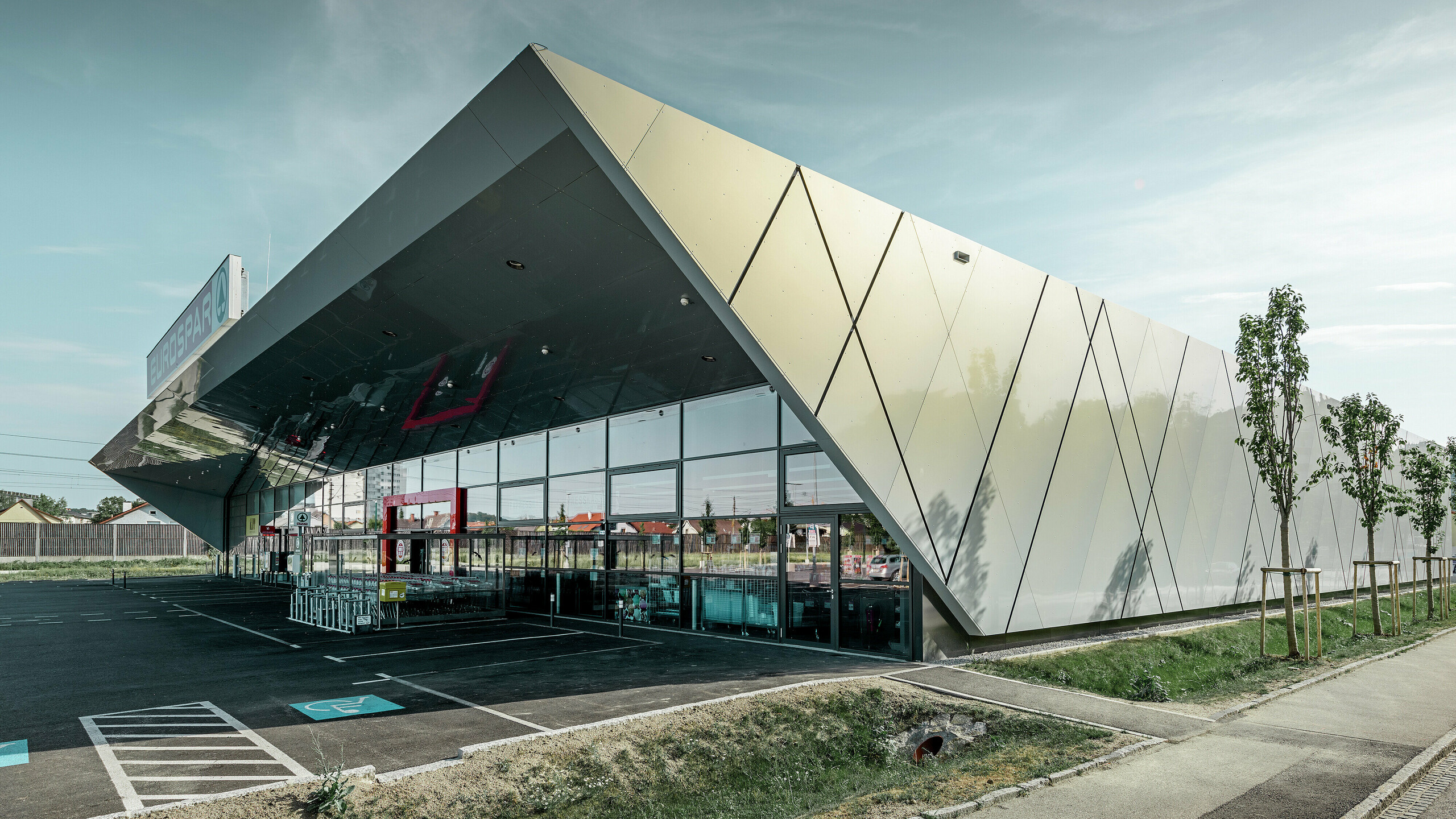 Entreegedeelte van het nieuwe Eurospar filiaal in Wels met het moderne gevelpaneel van PREFA in zilver goud; afhankelijk van de kijkhoek glinsteren de aluminium composietplaten in de zon in verschillende kleuren.