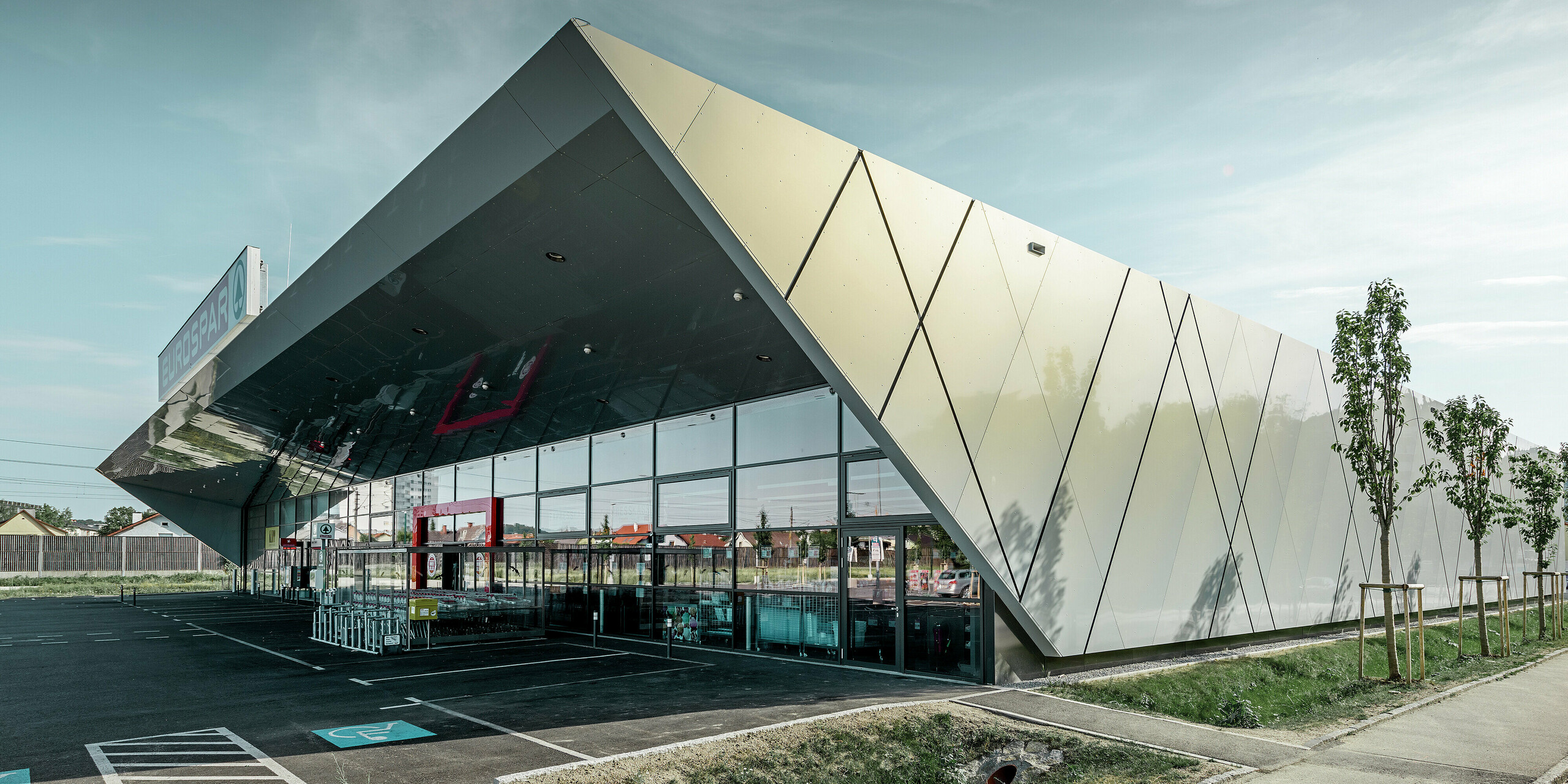 Entreegedeelte van het nieuwe Eurospar filiaal in Wels met het moderne gevelpaneel van PREFA in zilver goud; afhankelijk van de kijkhoek glinsteren de aluminium composietplaten in de zon in verschillende kleuren.