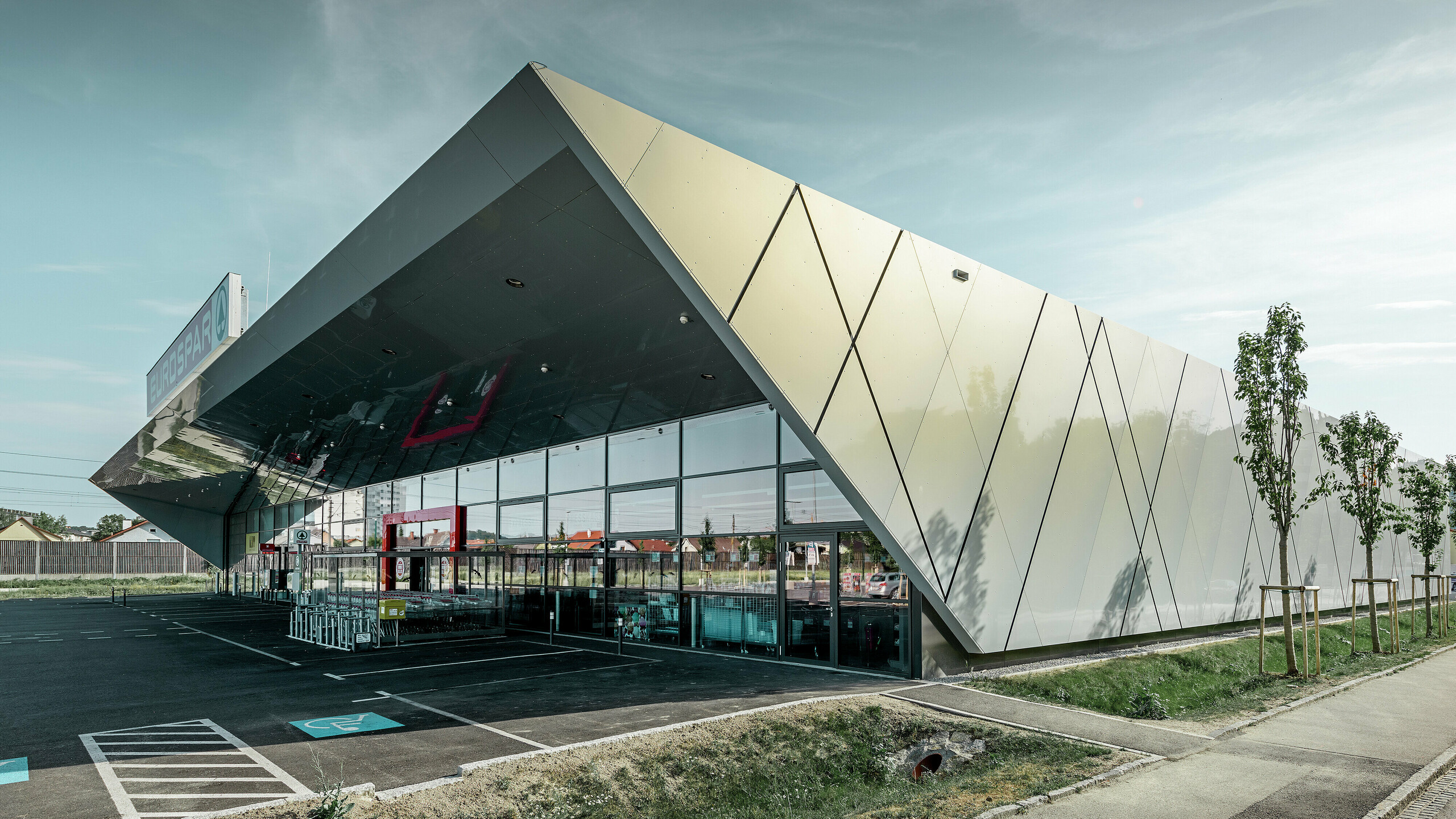 Entreegedeelte van het nieuwe Eurospar filiaal in Wels met het moderne gevelpaneel van PREFA in zilver goud; afhankelijk van de kijkhoek glinsteren de aluminium composietplaten in de zon in verschillende kleuren.