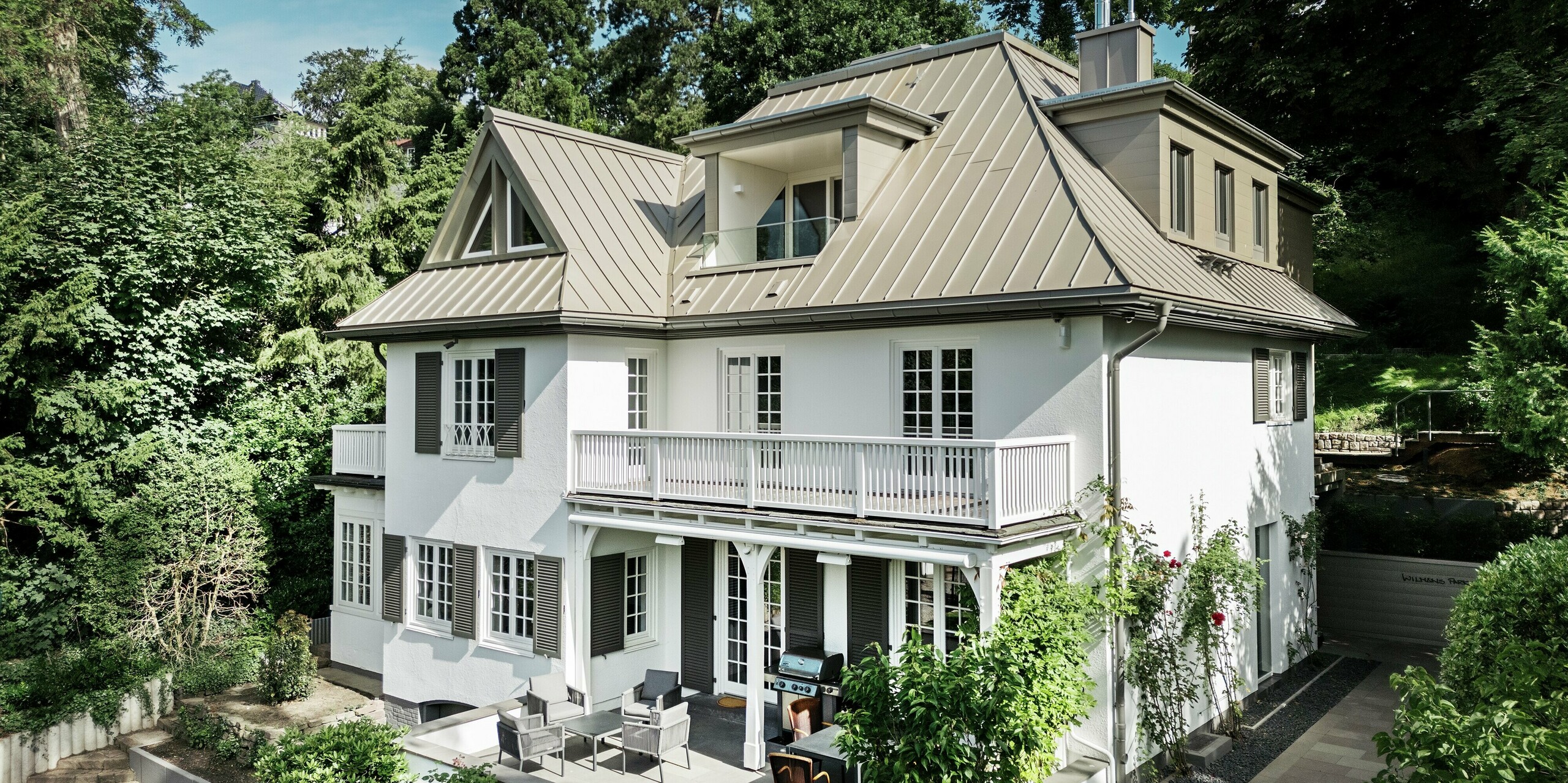 Das Bild zeigt die Villa Blankenese aus einer frontalen bzw. leicht schrägen Perspektive, eingebettet in eine grüne Umgebung. Das markante Dach aus PREFALZ in der Farbe Bronze prägt die Architektur und verleiht dem Gebäude eine elegante, moderne Note. Die Stehfalztechnik betont die klare Linienführung und sorgt für eine langlebige, witterungsbeständige Lösung. Ergänzend dazu sind an den Gauben und Fassadenteilen bronzefarbene PREFA Sidings eingesetzt, die das harmonische Gesamtbild abrunden. Die Kombination aus hochwertigen Aluminiumprodukten und klassischer Architektur macht die Villa zu einem stilvollen Referenzobjekt.