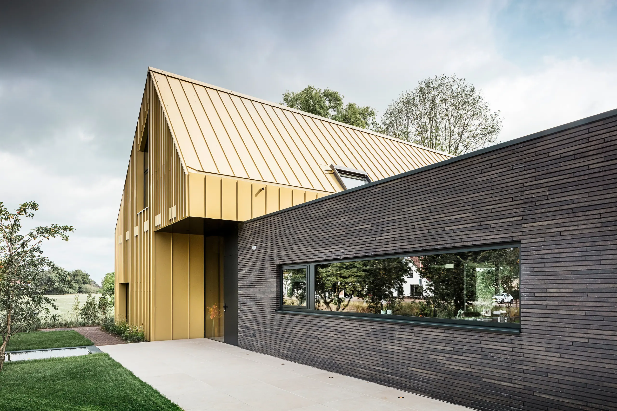 Modernes Einfamilienhaus mit flachem Anbau, das Hauptdach und Teile der Fassade sind mit PREFA Falzonal Savannenbeige Aluminium verkleidet; großes Panoramafenster im Anbau, umgebender Garten mit Rasen, Bäume im Hintergrund, bewölkter Himmel.