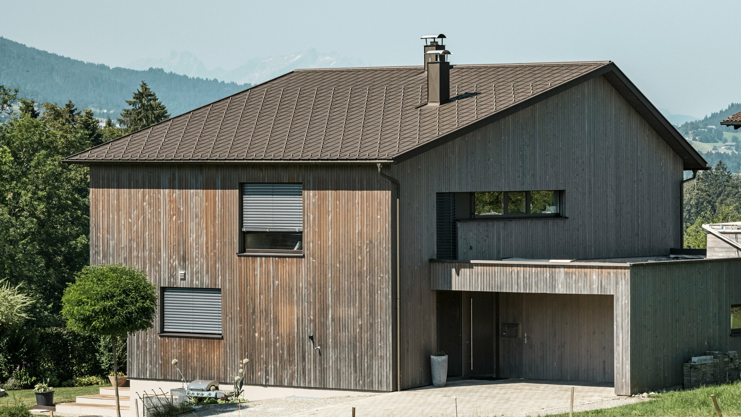 Zijaanzicht van een moderne vrijstaande woning in Krumbach met een combinatie van een gevel van natuurlijk hout en een duurzaam PREFA aluminium dak in P.10 bruin. De harmonieuze kleurstelling toont de elegante integratie van modern design en traditionele materialen, waarbij de PREFA- dakpannen voor een esthetisch en functioneel dakdesign zorgen.
