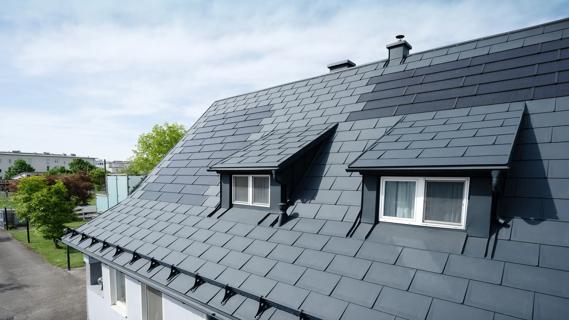 Detailfoto van een dak dat is bedekt met grote PREFA-solar-dakpannen en de PREFA-dakpan R.16 in P.10 antraciet. Er zijn twee dakkapellen te zien. De solar-dakpannen zijn gelijkmatig over het dak verdeeld en zorgen voor een moderne, duurzame oplossing.