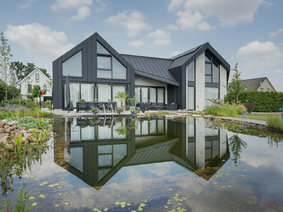 Idyllisches Bild eines Familienhauses in Veenendaal, Niederlande, das sich in einem klaren Gartenteich spiegelt. Das dunkle PREFALZ Dach- und Fassadensystem kontrastiert mit dem hellblauen Himmel, während der Teich das Haus und die umgebende Natur spiegelt und die friedliche Atmosphäre unterstreicht. Ein gepflegter Garten mit einer Vielzahl von Pflanzen und Sitzgelegenheiten lädt zum Verweilen ein und vervollständigt das Bild einer harmonischen Wohnästhetik in Verbindung mit der Natur.
